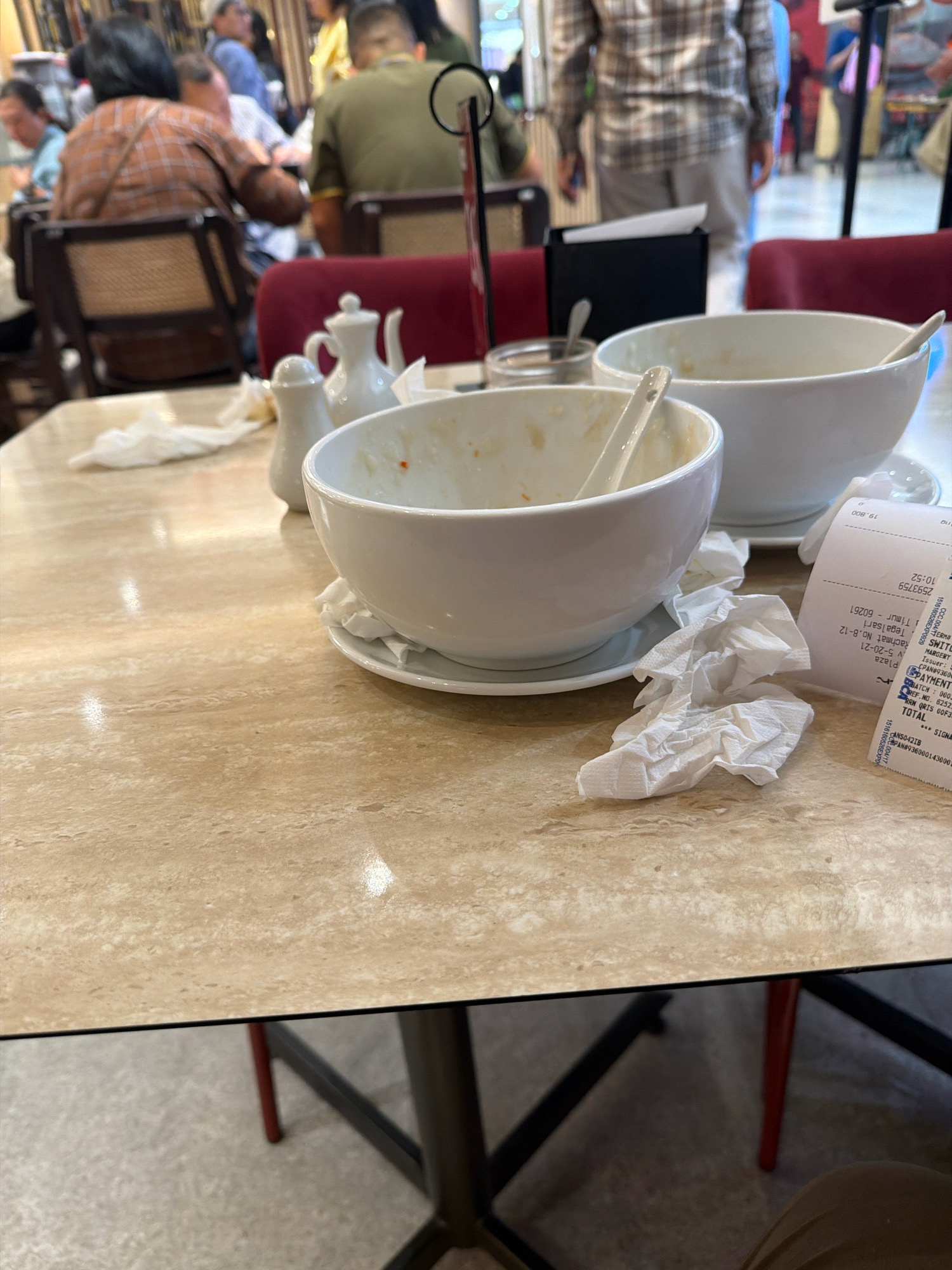 The image shows a close-up view of a table in a restaurant setting, likely in a city like Unknown, Unknown. On the table, there are two empty white bowls, one larger than the other, with spoons in them. A white salt and pepper shaker set and a crumpled white napkin are also present. To the right of the bowls, a receipt is visible. In the background, blurred figures of people can be seen sitting at tables and walking around. The table surface appears to be a light-colored marble or laminate. The overall scene suggests that a meal has recently concluded.