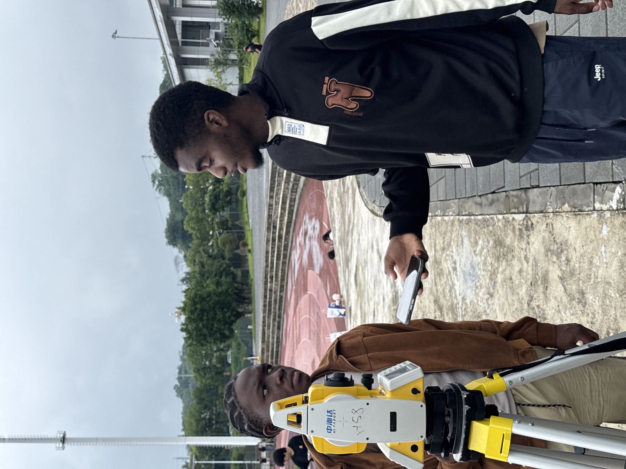 In Jiangjin, China, two young Black men are standing on a paved area next to a running track. The man on the left, wearing a brown jacket over a tan shirt and khaki shorts, is positioned next to a yellow and white surveying instrument mounted on a tripod. The instrument has Chinese characters and "HI-TARGET" written on it. The man on the right, wearing a black quarter-zip sweatshirt with white and cream accents and navy blue sweatpants, is holding a silver smartphone. He appears to be showing the phone to the man on the left, who is looking at the phone. The background shows a running track, bleachers, trees, and a modern building. The sky is overcast.