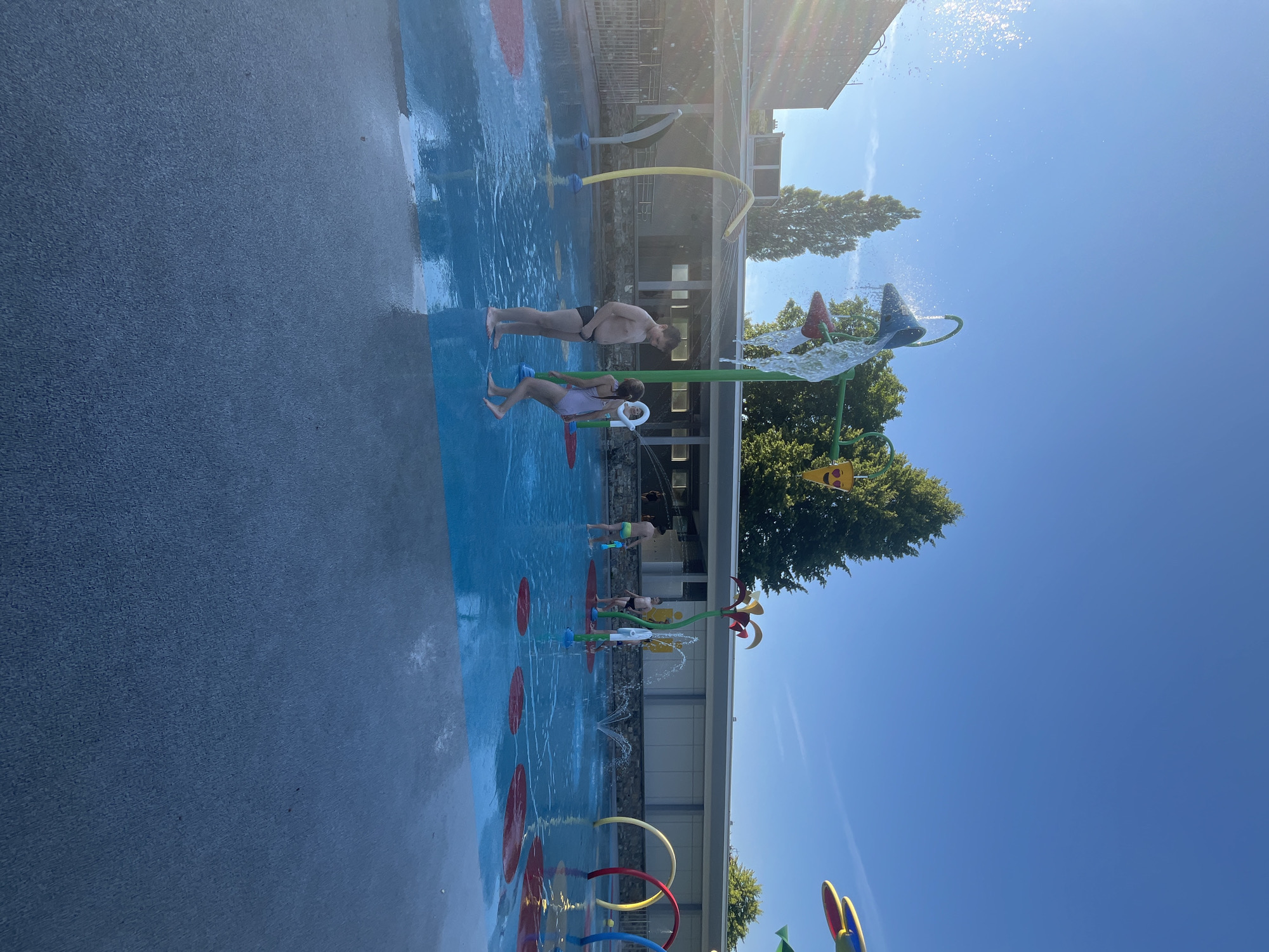The image depicts a sunny day at a splash pad, a water play area for children. The scene is populated with several people, including adults and children, enjoying the water features. In the foreground, a man stands with his back to the camera, while a woman in a pink swimsuit is positioned next to him, seemingly interacting with a water spray. Further back, other individuals are visible, some standing in the water and others near the edge of the splash pad.
The splash pad itself features blue ground with red circular designs. Various water play structures are present, including colorful arches and spray elements that are actively spraying water. In the background, a building with windows and a roof is visible, suggesting it might be a changing facility or a part of a larger recreational complex. Lush green trees are also present, framing the scene and indicating an outdoor setting. The bright blue sky and the presence of sun flares suggest that it is a clear and sunny day. The overall mood appears to be cheerful and playful, with people engaged in recreational activities. No visible text is present in the image.