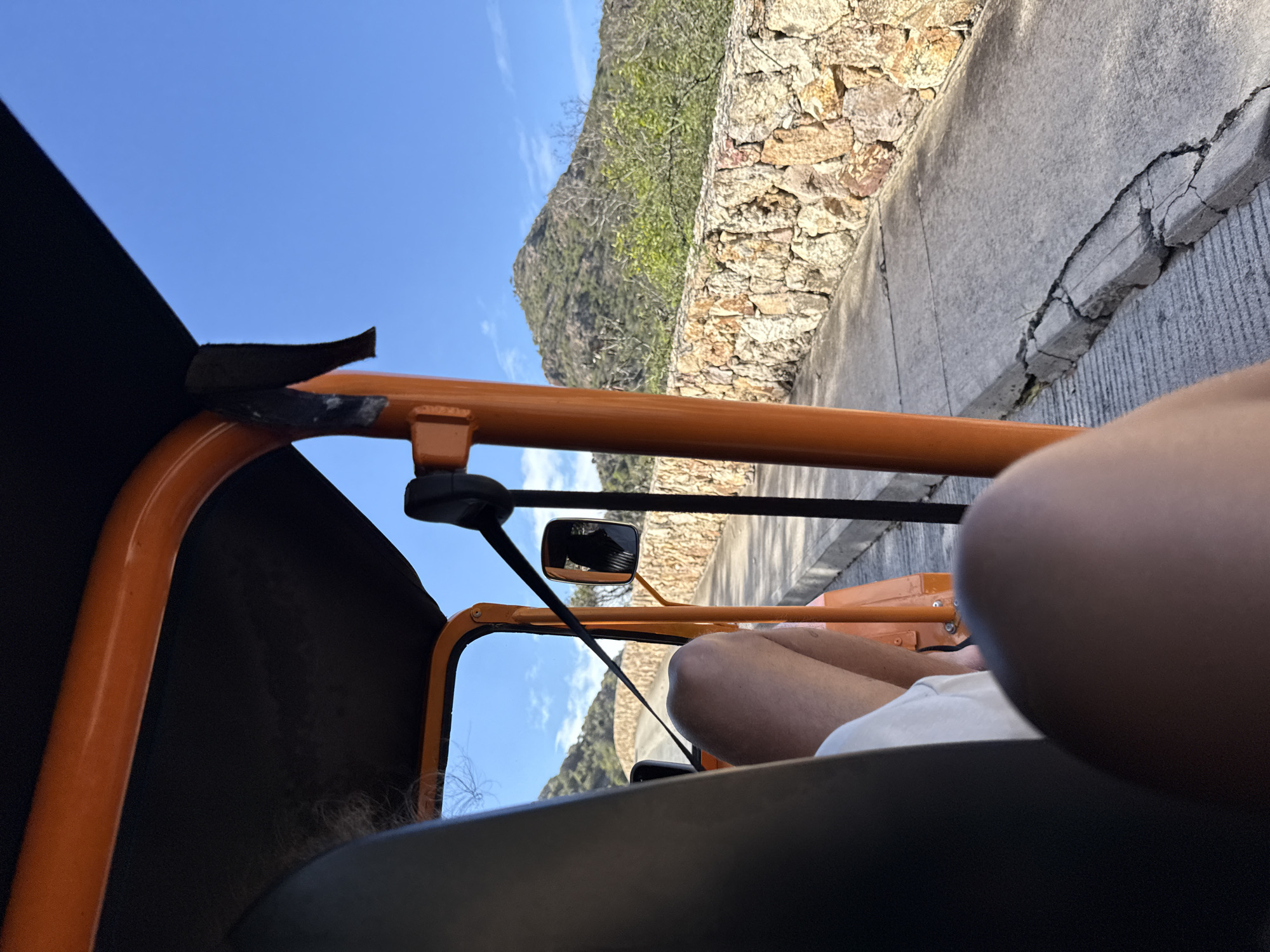 The image, taken from inside an orange, open-air vehicle, likely a taxi or buggy, shows a ride along a road in Saint Barthélemy, France. The camera angle suggests the photographer is a passenger. The road has a cracked concrete edge and a stone wall. In the background, a mountain covered with vegetation is visible under a clear blue sky. Within the vehicle's frame, there's an adjustable side mirror. Part of the passenger’s leg and a white garment are in the foreground, obscuring part of the vehicle's interior. The black canvas roof of the vehicle provides a partial shade.