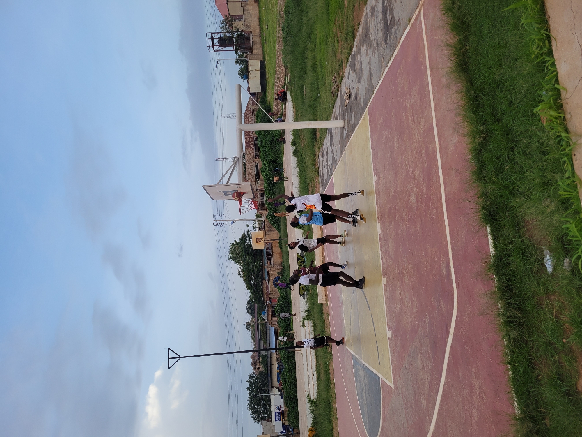 This media file depicts a group of young people on an outdoor basketball court. The scene is viewed from a high angle, looking down onto the court. The court itself has a red and yellow painted surface with white lines. To the right of the court is a strip of lush green grass. In the background, there are buildings, trees, and a water tower, suggesting an urban or semi-urban setting. The sky is overcast with a light blue hue, indicating it might be daytime, possibly late afternoon.
Several individuals are present on the court. In the foreground, two people are engaged in a basketball game, with one attempting a shot at the hoop while the other defends. A basketball is visible near the hoop. Further down the court, other individuals appear to be observing or waiting their turn. They are dressed in casual athletic wear. The overall mood seems relaxed and recreational, with a focus on the sport.
There is some visible text on the clothing of one of the individuals on the court, which appears to say "ELITE". The overall setting and activity suggest a casual pick-up basketball game or practice session. The weather appears to be mild and possibly humid, given the overcast sky.