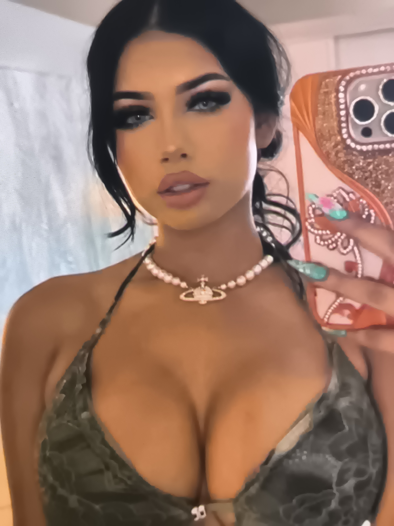 The image shows a close-up of a young woman taking a selfie. She has dark hair styled in loose waves and is wearing full makeup, including dramatic eyeliner and false eyelashes. She has bright blue eyes and full lips. Around her neck, she wears a pearl necklace with a pendant. Her attire appears to be a dark-colored bikini top with a textured pattern.
In her right hand, she holds a phone with an orange, glittery case adorned with decorative elements and a large camera bump. Her fingernails are long and painted in a vibrant teal color with additional designs on some.
The background is slightly blurred but suggests an indoor setting, possibly a bathroom or bedroom, with hints of a textured wall. The lighting appears to be artificial, casting a warm glow on her skin. The overall mood seems to be one of casual vanity and self-expression. There is no visible text in the image.