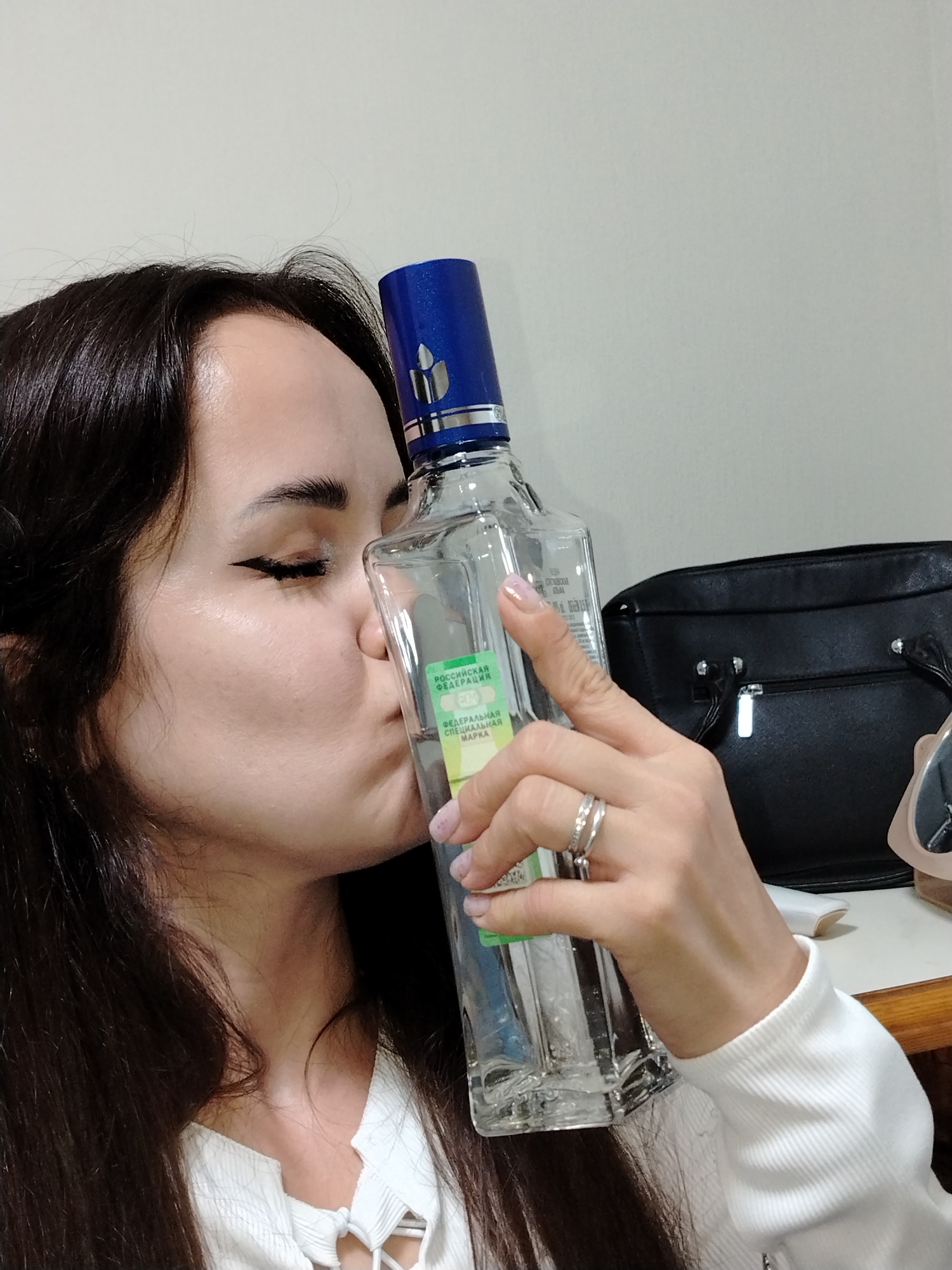 A woman with dark hair and eyeliner is shown in an indoor setting, identified as being in the city of Chaikovsky, Russia. She is depicted kissing a clear glass bottle of "Moskovskaya" vodka, recognizable by its green and white label displaying "РОССИЙСКАЯ ФЕДЕРАЦИЯ" (Russian Federation) and "ФЕДЕРАЛЬНАЯ СПЕЦИАЛЬНАЯ МАРКА" (Federal Special Mark), and a blue cap featuring a candle flame logo. Her eyes are closed as her lips press against the bottle's side. She wears a white top with a laced V-neck and a white long-sleeved ribbed garment. Two rings are visible on her left ring finger, and her fingernails have light pink polish. In the background, a black handbag with a silver zipper and part of a lighter-colored object are visible on a wooden surface, against a plain, light-colored wall.