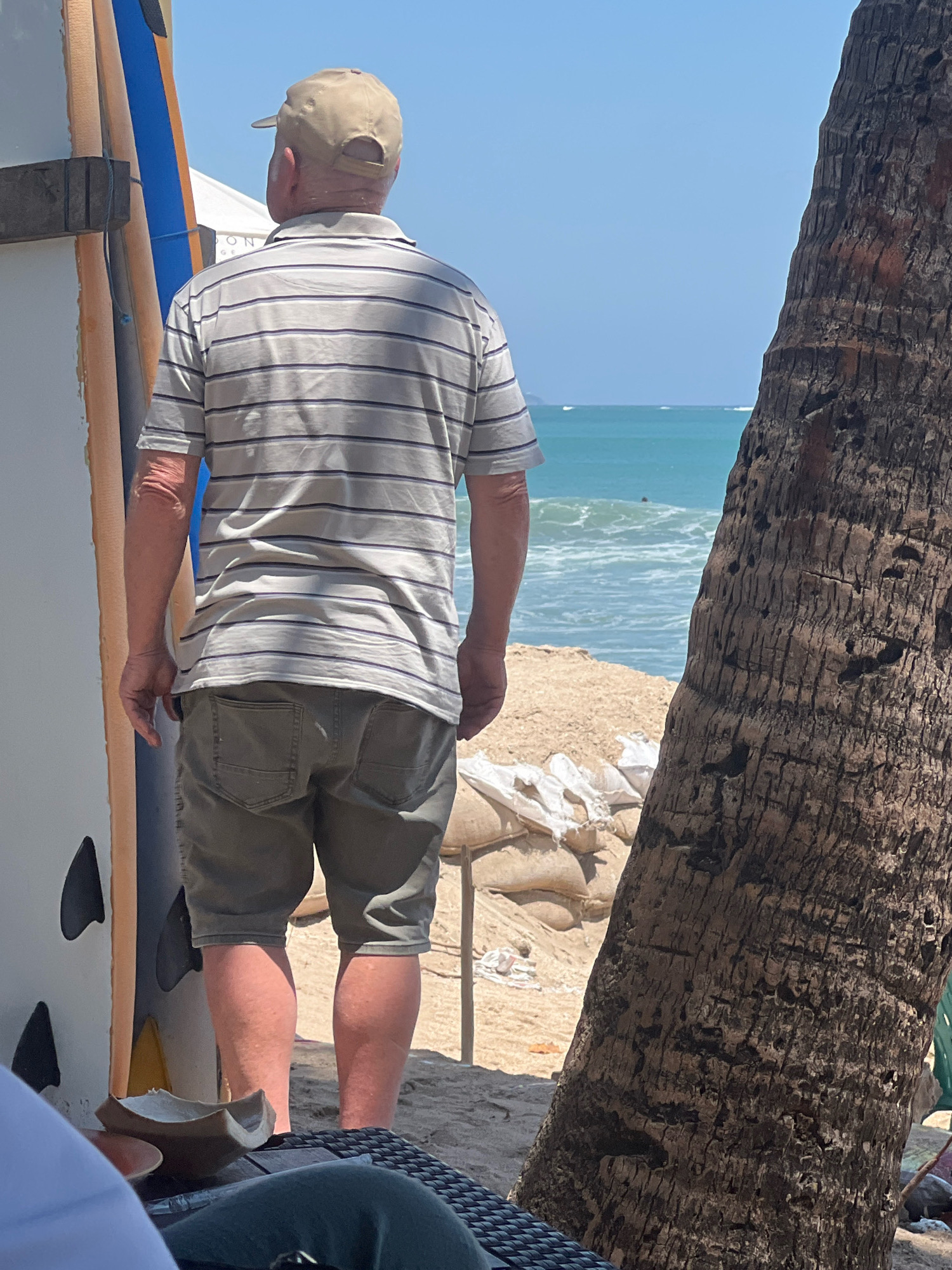 An overhead shot shows a man in a light-colored, horizontally striped polo shirt and khaki shorts standing on a sandy beach in Kuta, Indonesia. He is wearing a tan baseball cap and looking out at the ocean. To his left are several surfboards propped up against a white structure. To his right is the textured trunk of a palm tree. In the background, sandbags are piled on the beach, and the turquoise ocean stretches to the horizon under a clear blue sky. The man's left arm is visible, showing some sun damage. The scene appears to be a bright, sunny day, likely during tourist season.