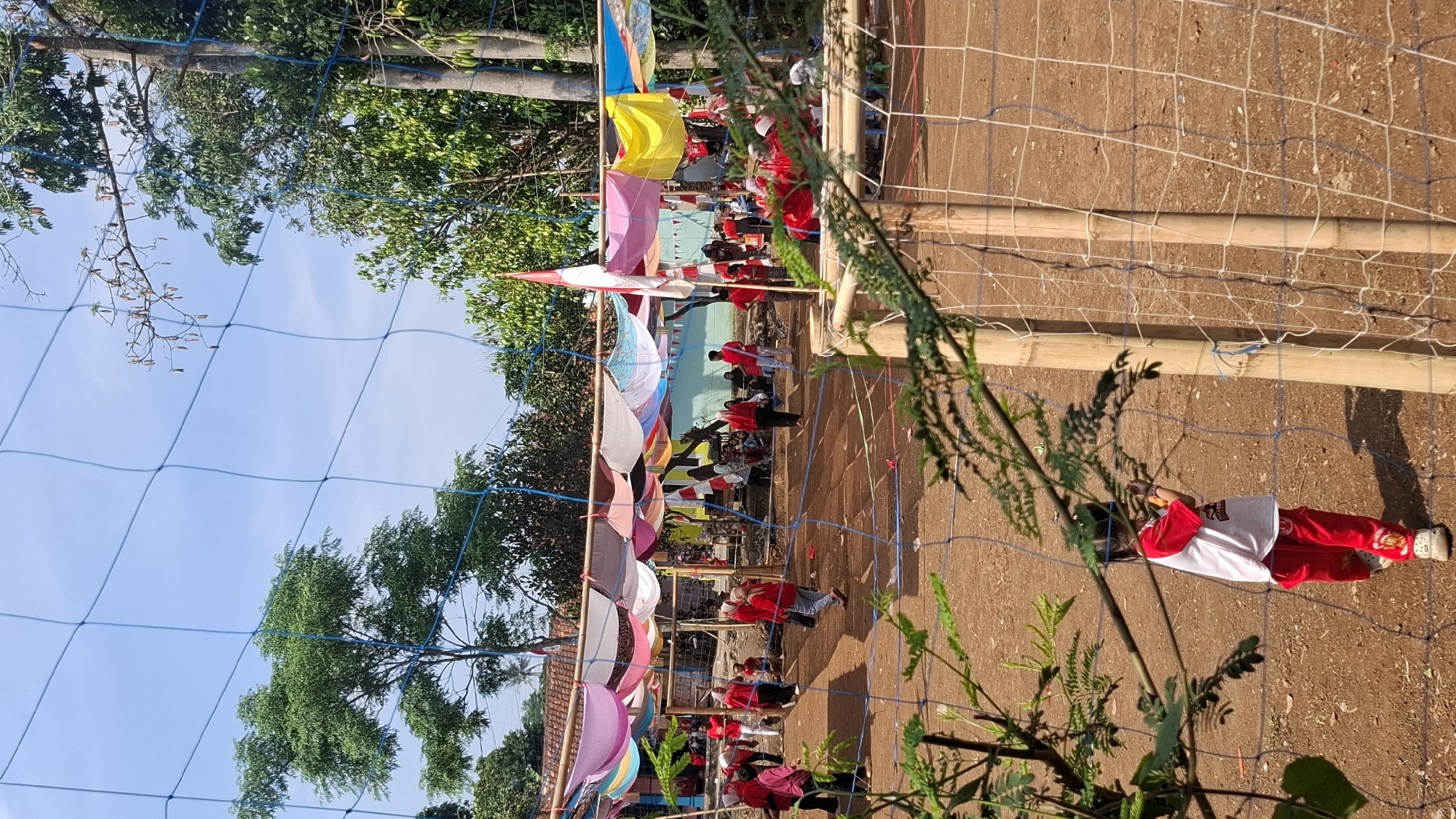 The image depicts an outdoor scene, likely in Cilaku, Indonesia, during a daytime event. In the foreground, a child wearing a red and white shirt and red pants stands facing away from the camera, partially obscured by a net and some green foliage. The child appears to be interacting with the net.
The background reveals a gathering of people, many of whom are wearing red clothing. They are positioned under a makeshift canopy supported by bamboo poles. The canopy is adorned with colorful fabric draped overhead, creating a festive atmosphere. Several flags, including what appears to be an Indonesian flag, are visible. The ground is a dirt surface, suggesting an open field or court. Trees are present around the perimeter of the area. The sky is blue and clear. The overall scene suggests a community event or celebration.