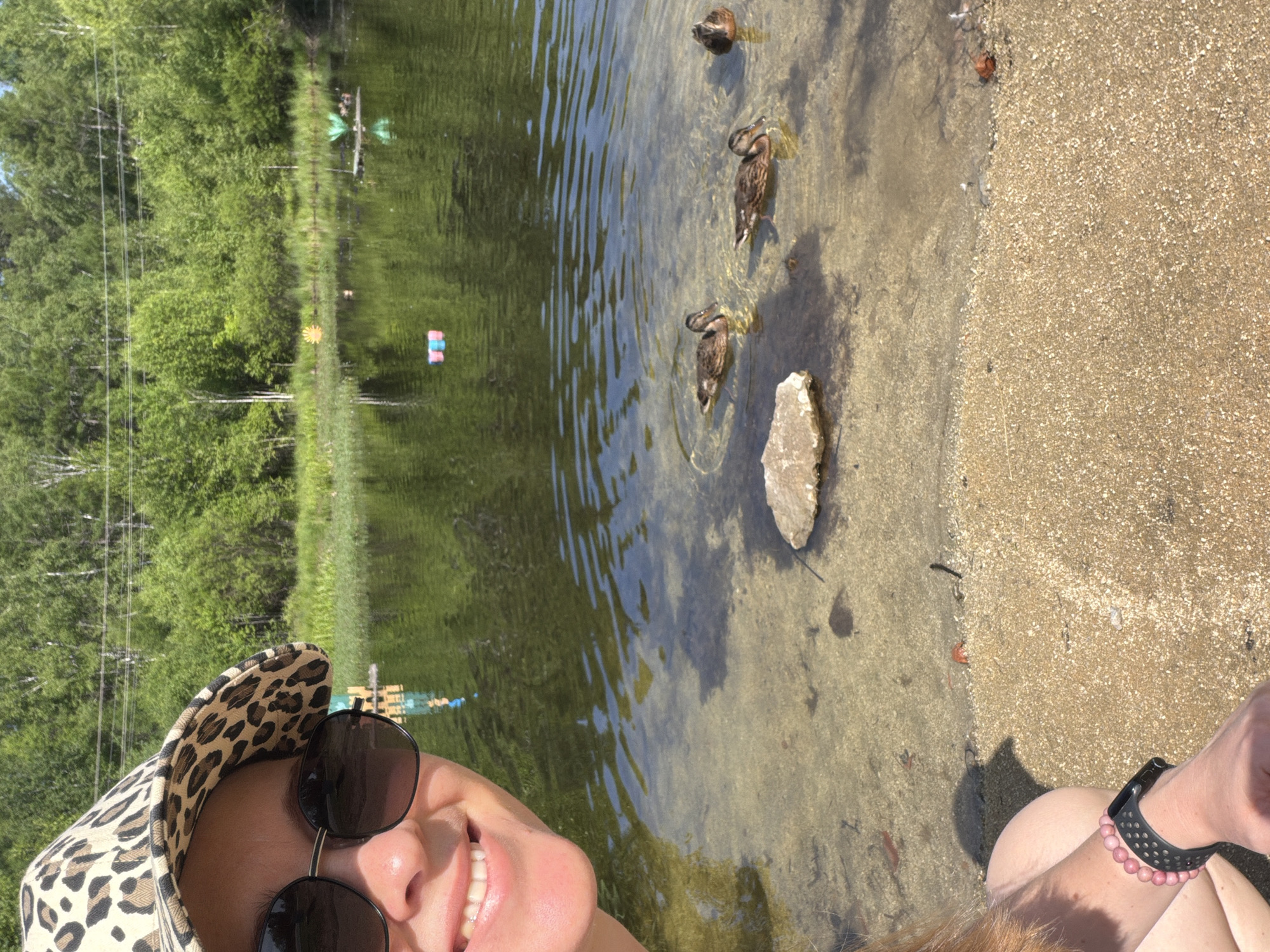 The image is a selfie taken by a person wearing a leopard-print hat and sunglasses, smiling at the camera. They are sitting by the edge of a clear, shallow body of water with a sandy bottom. Several ducks are swimming in the water in the foreground. In the background, there is a lush green forest and a calm body of water with some colorful objects floating on it. The image depicts a sunny day in a natural outdoor setting, likely a park or a recreational area. The location of Кульдур, Russia is not evident from the visual information provided in the image.