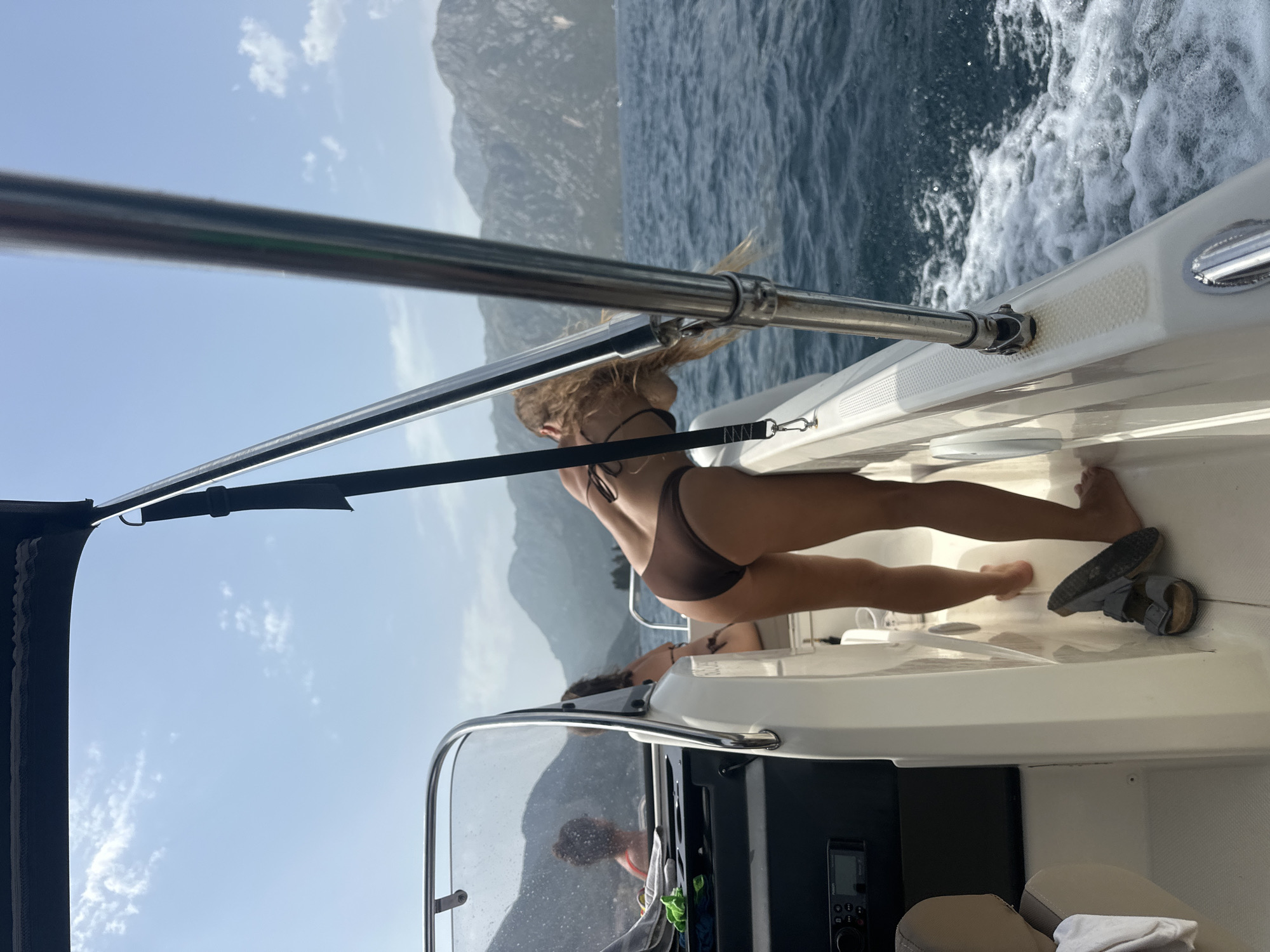 An outdoor, medium shot captures a woman in a brown bikini on a boat sailing on the waters near Perast, Montenegro. The woman, seen from the rear, bends over the railing with her blonde hair falling forward. She is positioned on the right side of the boat, her legs bent as she leans. The boat is moving, creating a wake of white foam in the choppy blue water. In the background, large, rugged mountains rise from the water under a hazy sky with scattered white clouds. The interior of the boat is visible on the left, with seating and a dashboard. Another person, with dark curly hair, is seated further back in the boat, facing away from the camera and towards the mountains. The overall scene depicts a leisurely boat trip on a sunny day.