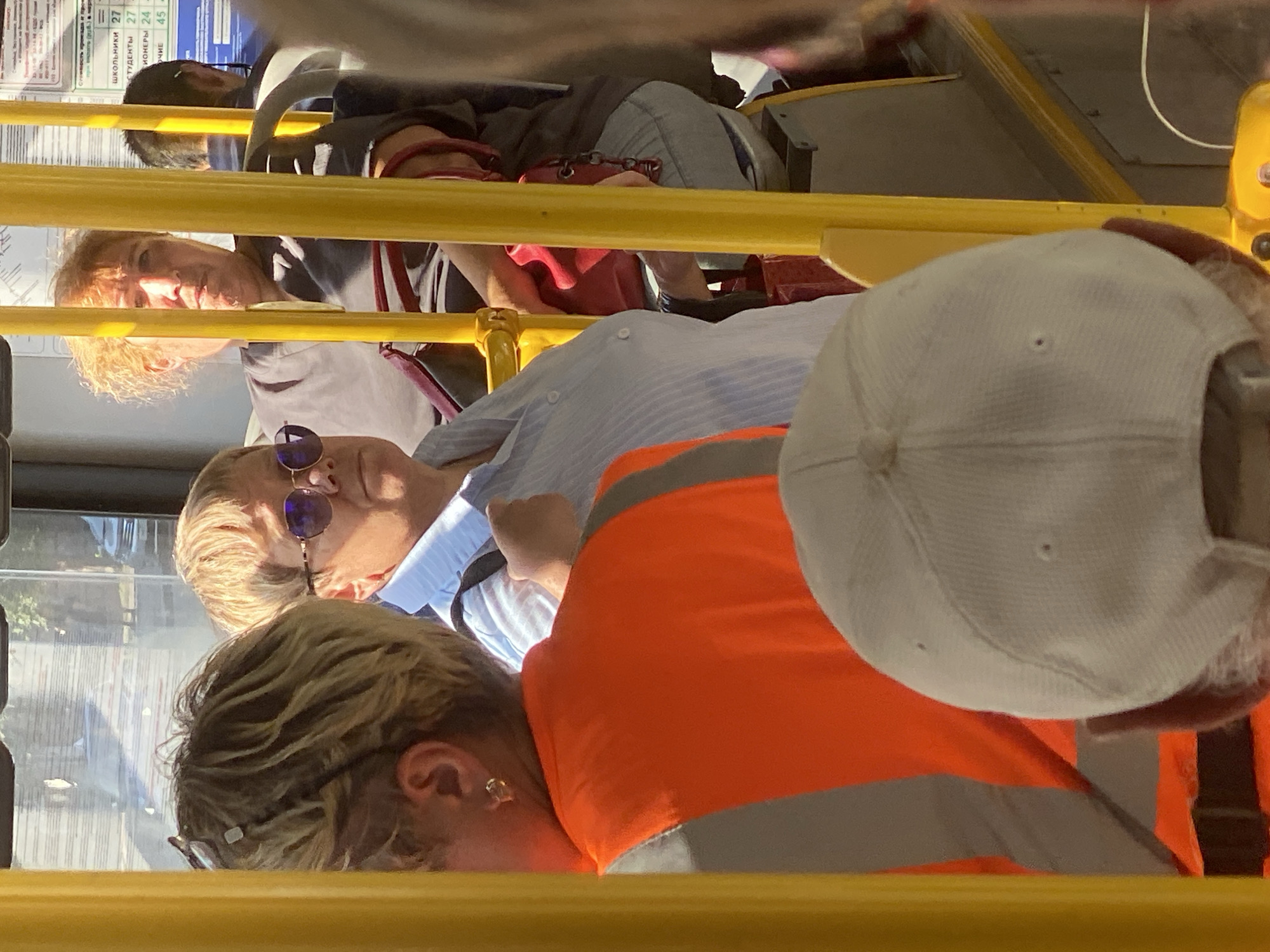 The image shows the interior of a public transport vehicle, likely a bus, with several passengers. In the foreground, a person wearing a white baseball cap is visible from the back. To their left, another person is seen from the side, wearing a light blue striped shirt and sunglasses, with blonde hair. Further back and to the right, a person with curly blonde hair is looking forward. Yellow poles are prominent throughout the scene, a common feature in public transport. The environment suggests an urban setting, and based on the presence of Cyrillic text on a sign in the background, the location is likely Novokuznetsk, Russia. The sign appears to display route or schedule information, with some numbers and text visible, although not fully legible.
