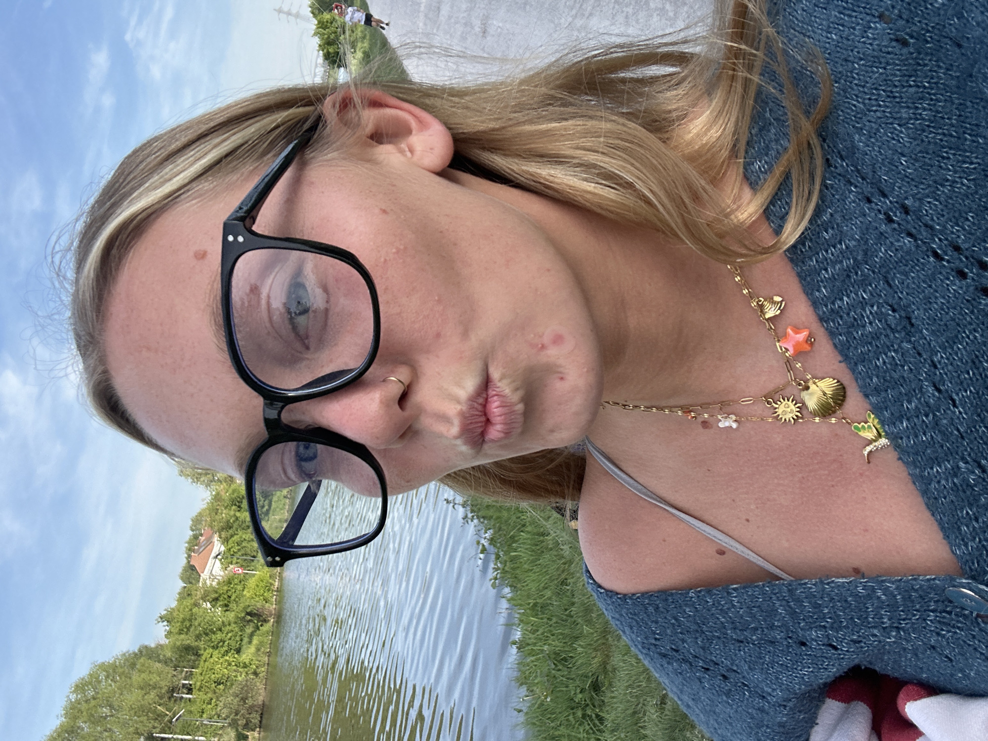 A woman is shown in a close-up, outdoor shot. She is wearing black glasses, a blue sweater, and a gold necklace with charms. She has blonde hair and is making a kissing face. In the background, there is water, grass, trees, and a building. In the distance, two people are walking on a path. The photo was likely taken in Marquette-lez-Lille, France, given the presence of typical architecture of the region.