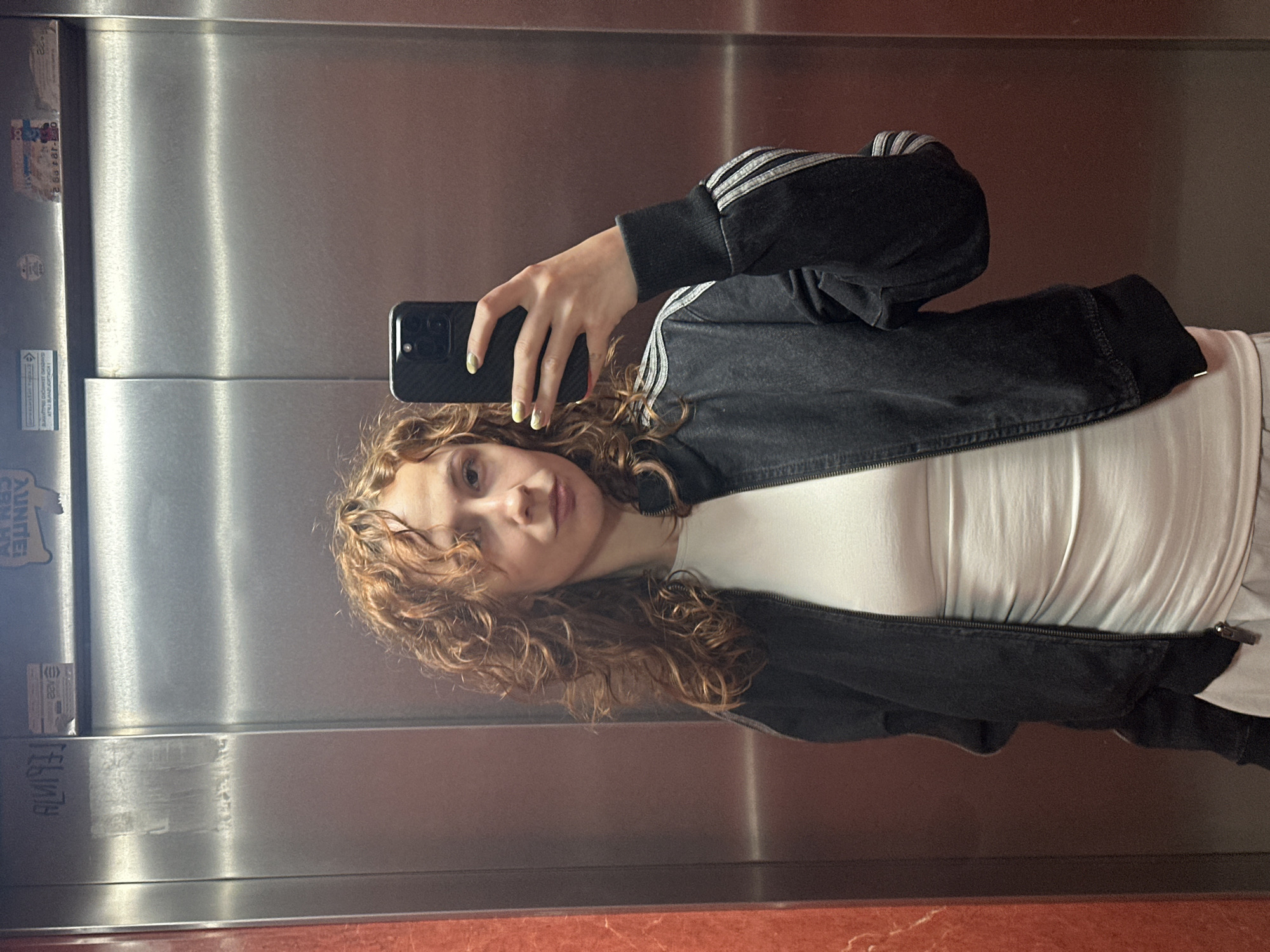 A young woman with curly, reddish-brown hair is captured in a selfie taken in a reflective surface, likely a polished metal elevator wall, in Novi Sad, Serbia. She holds a dark-colored smartphone with a textured case, angled towards the reflection. Her nails are painted a light yellow or neon green. She is wearing a black jacket with distinctive white three-stripe detailing on the sleeves, characteristic of Adidas, over a plain white top.
The setting appears to be an indoor space, possibly an elevator, characterized by a tall, reflective, grayish-silver metal wall that extends vertically, divided by a central seam. The bottom edge of the reflection shows a reddish-brown, possibly marble or tiled, surface.
Visible on the left side of the metal wall are several stickers and some graffiti. One sticker shows partial text "AUNDE" and "UN," along with a phone number fragment "08:18 99 2". Below it, a smaller rectangular sticker has faint text including "18/22". Further down, inverted text, possibly "FELIPE" or "FELIVY," is scrawled vertically on the wall. The woman's expression is neutral, and her gaze is directed towards the camera.