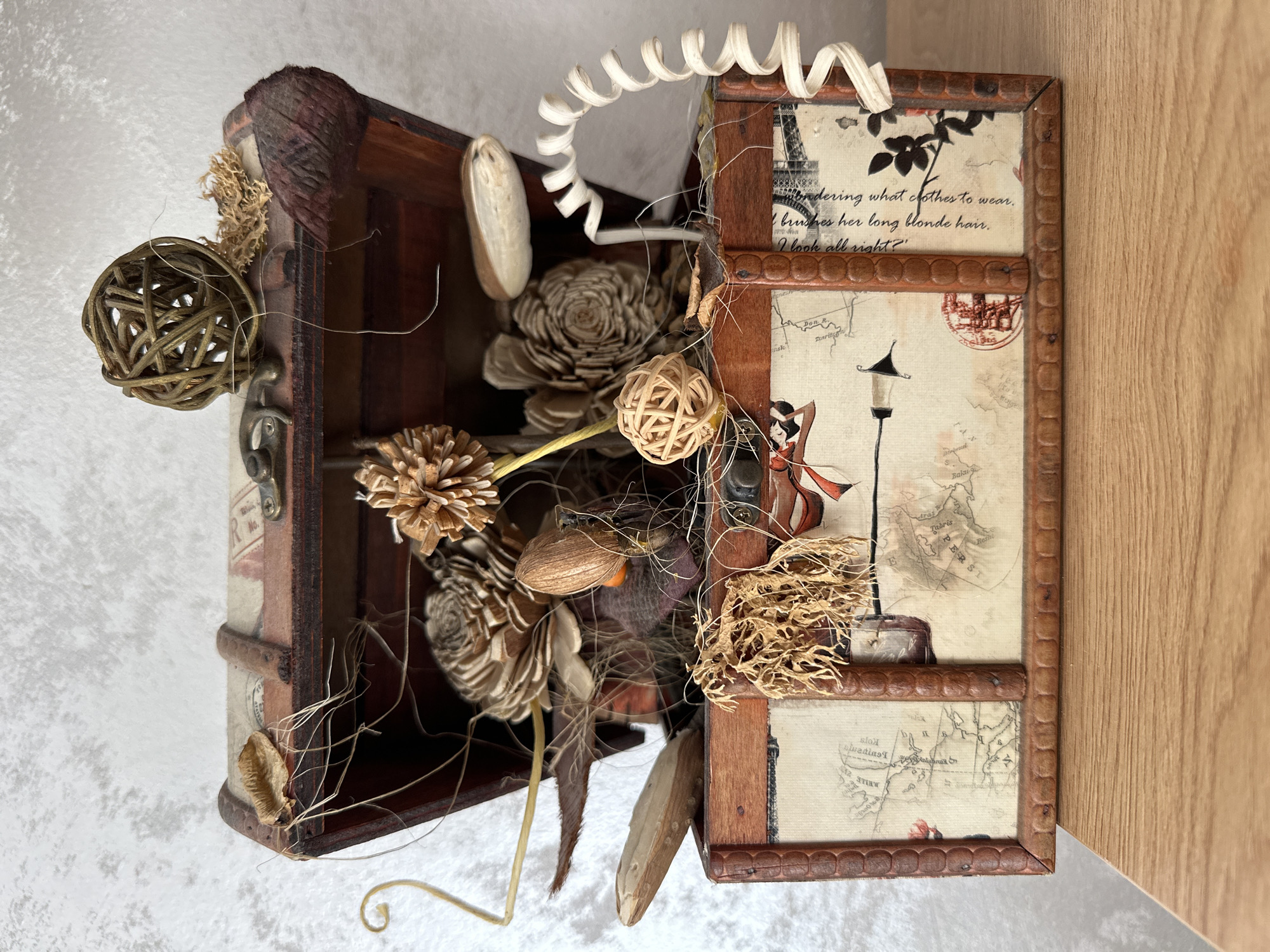 The image displays a decorative, open wooden chest filled with various natural and craft items. The chest has a vintage aesthetic, with faux leather trim and straps, and the front panel is decorated with a collage of map fragments, script, and imagery. Inside, the chest contains items such as dried flowers, pinecones, small woven balls, and other organic materials arranged artfully. A coiled white ribbon-like material extends from the right side of the chest. The chest rests on a light-colored wooden surface, set against a textured, light gray background. There are no people or animals visible in the image. The scene does not depict any recognizable location in Glavatartsi, Bulgaria, or any other specific geographical area. The context provided regarding Glavatartsi, Bulgaria, is not supported by the visual information in the image.