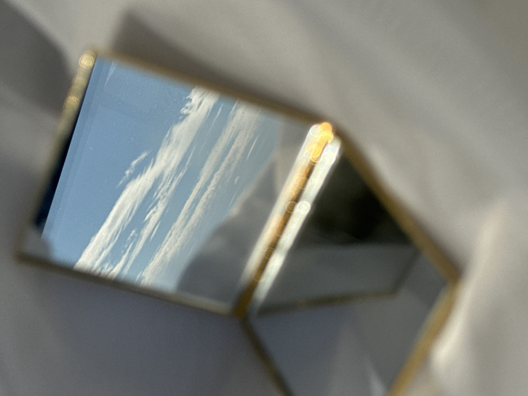 The image shows a small, geometric, glass box with a gold trim. The box is reflecting a bright blue sky with some white clouds. The reflection appears to be taking place inside, as a white surface is seen underneath the box and out of focus. The location is unknown.