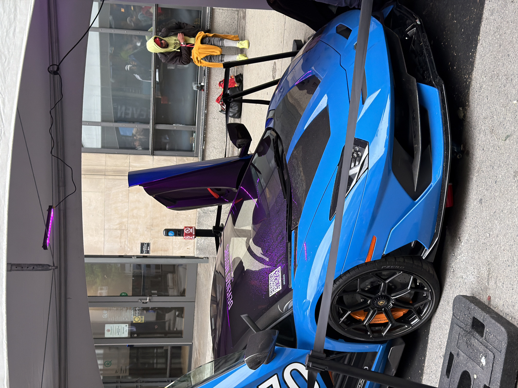 The image shows a blue and purple Lamborghini with an open door, parked on a city street in front of a building with glass doors and windows. A person wearing a yellow hoodie and jacket is lying on the sidewalk next to the car. A parking meter is visible near the car, and the reflection in the building window suggests the location is Montreal, Canada, with the word "EVENT" visible in the window's reflection. There is also a purple light on the top left corner. The car is surrounded by a black barricade with yellow strip.