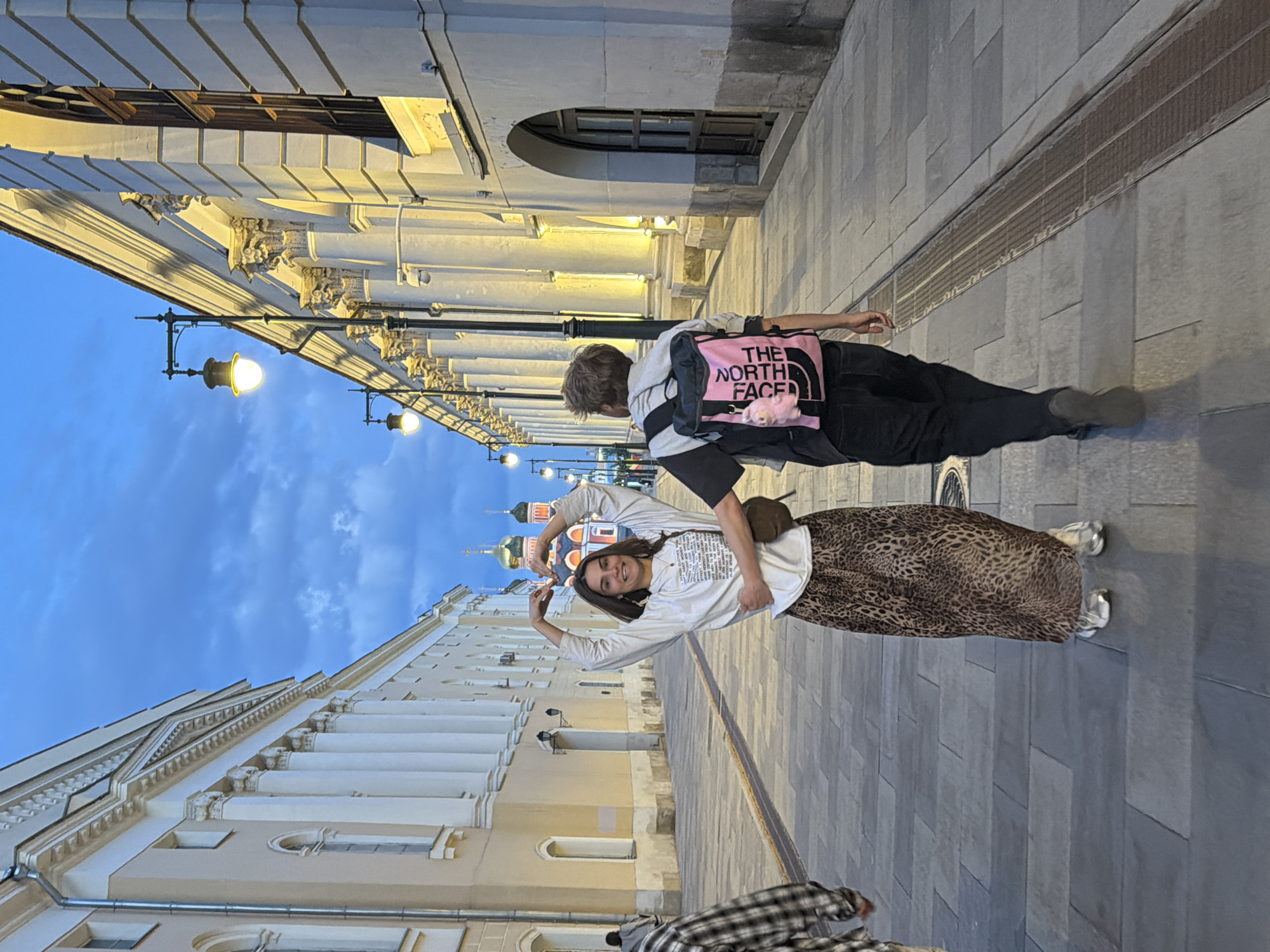 The image depicts two individuals on a paved street in an urban setting at dusk or early evening. On the right, a female-presenting person with long brown hair smiles directly at the camera, forming a heart shape with her hands. She is dressed in a white long-sleeve shirt featuring black text, a brown and black leopard print maxi skirt, and silver shoes. Beside and slightly behind her, a male-presenting person with short brown hair stands with an arm around her, facing away from the camera. He wears a grey hoodie, black trousers, and a pink and black shoulder bag with "THE NORTH FACE" clearly visible on its side.
The street is paved with rectangular grey tiles, and a long drainage grate runs alongside the buildings. The background features grand, multi-story buildings in a classical architectural style, with ornate columns, archways, and detailed cornices illuminated by artificial lights. Streetlights are also on, casting warm light onto the scene. The sky above is a deep blue with hints of clouds, consistent with twilight. In the distance, distinctive domed structures, reminiscent of onion domes, are visible, strongly suggesting a location in Moscow, Russia. The mood conveyed is one of joy and affection.