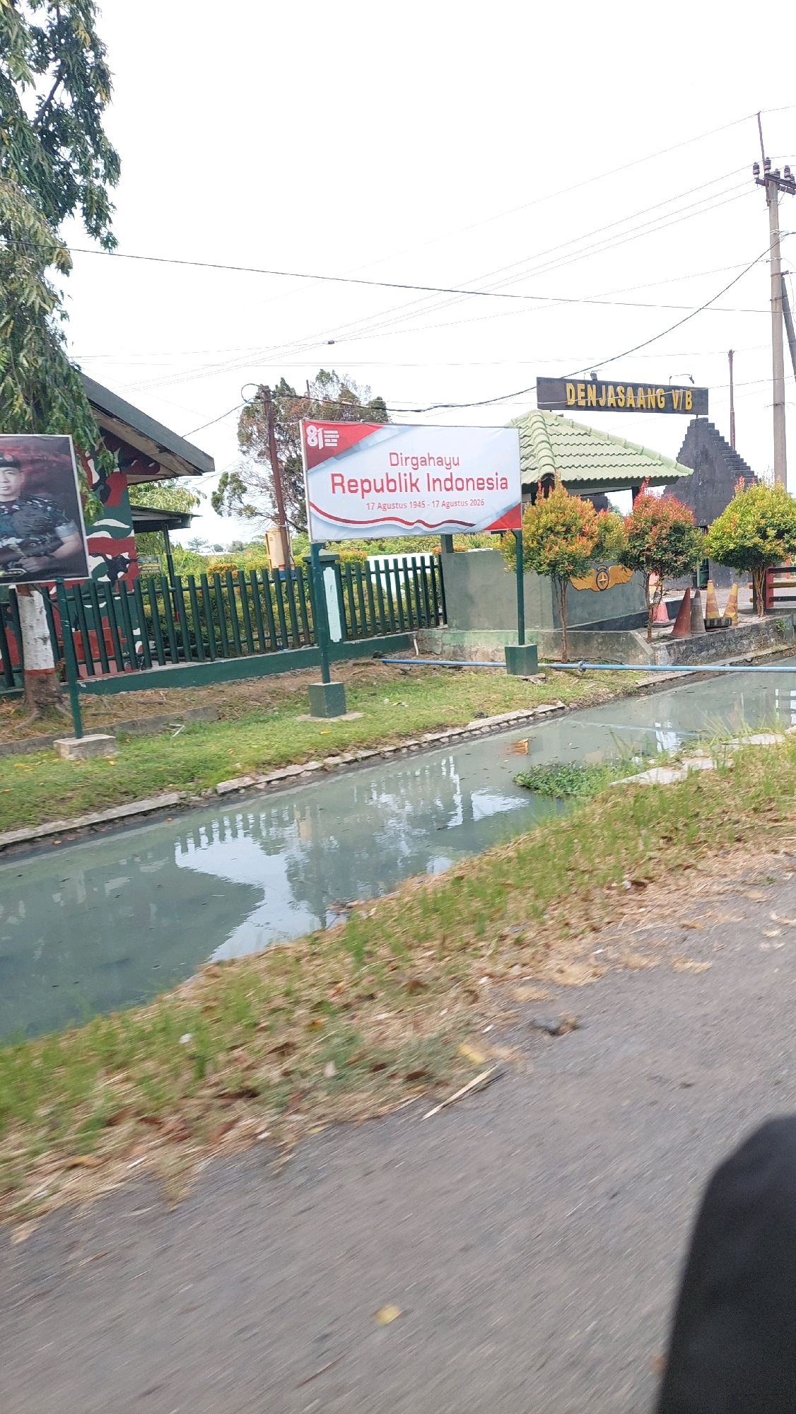 The image depicts an outdoor scene, likely in the city of Ketimang, Indonesia. In the foreground, a paved road runs diagonally across the lower portion of the frame. To the left of the road, a narrow, murky canal with some green vegetation growing in it is visible. Beyond the canal, a grassy area is present, leading to a dark green fence.
Behind the fence, a large white banner is prominently displayed, featuring red text that reads "Dirgahayu Republik Indonesia" (Long Live the Republic of Indonesia). Below this, smaller text indicates "17 Agustus 1945 - 17 Agustus 2026". To the left of the banner, a portrait of a man in military uniform is mounted on a camouflaged wall.
Further back, a building with a green, tiled roof is visible, and above it, a dark sign reads "DEN JASAANG VIB". Several small trees with orange and green foliage are planted in front of the building. In the background, utility poles and wires are present against a bright sky. The overall impression is of a public space or a facility decorated for a national celebration.