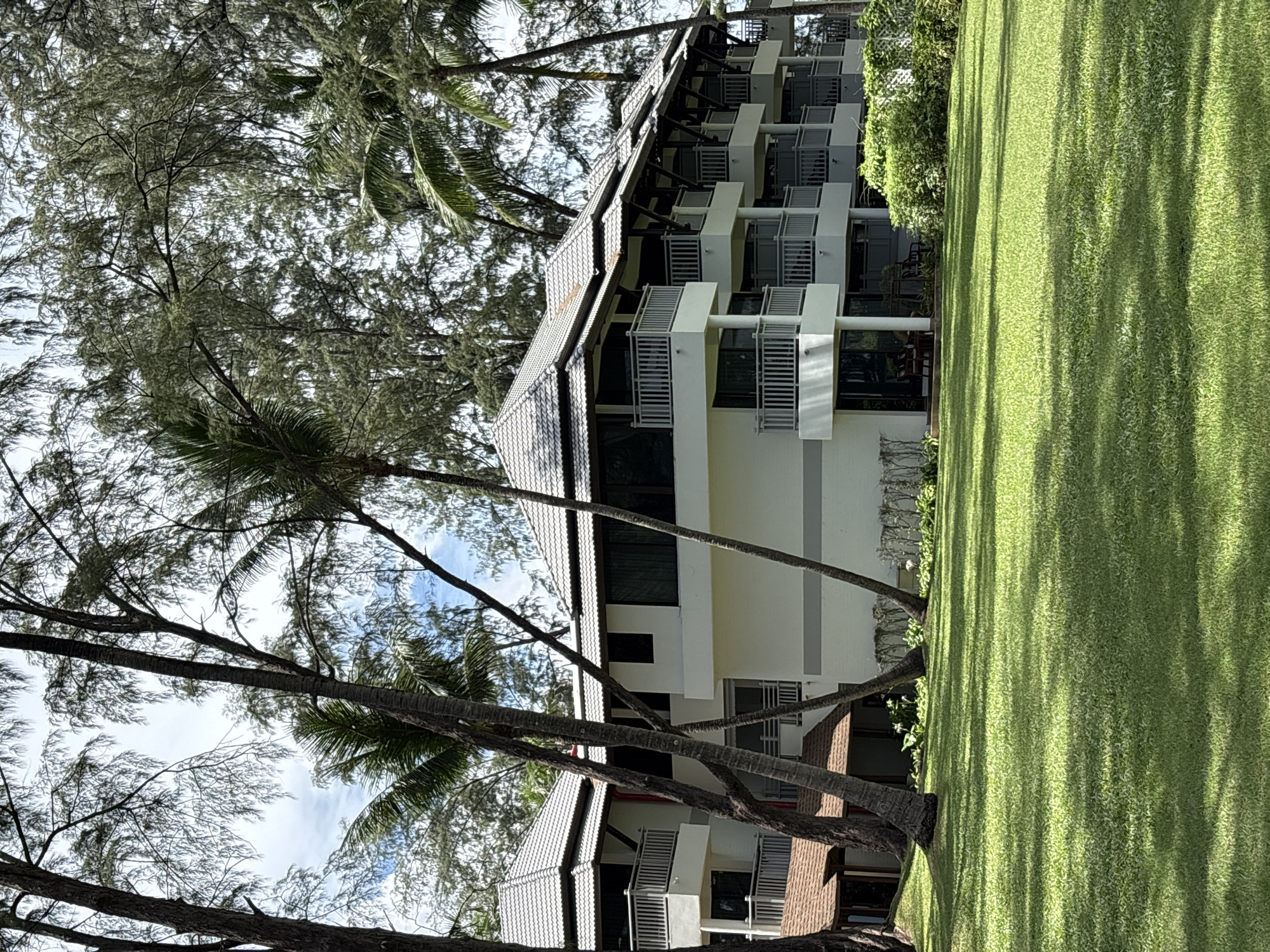 The image shows a multi-story building with a tiled roof and balconies, surrounded by lush green grass and tall trees, including palm trees. The building appears to be a hotel or resort, with a clean, modern design. The architecture suggests a tropical location. The sky is partly cloudy. The scene conveys a sense of tranquility and vacation. This building is located in Choeng Thale, Thailand.