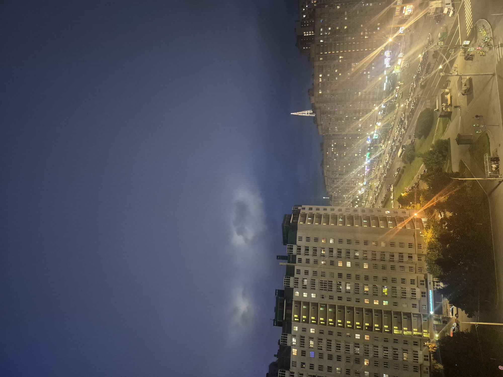 The image captures a nighttime view of a residential area in Saint Petersburg, Russia, likely a district with high-rise apartment buildings. The sky is dark and cloudy, with some lighter areas suggesting distant clouds or perhaps faint lightning. In the foreground, a large, multi-story apartment building is brightly lit from within by numerous yellow and some blue lights visible through its many windows. The building features a distinctive architectural style with prominent balconies and a dark-colored upper section.
To the right of the foreground building, a wide street is visible, lined with more apartment buildings that are also illuminated, creating a bright, shimmering effect due to streetlights and illuminated windows. A significant traffic jam is apparent on the street, with many cars queuing bumper-to-bumper, their headlights and taillights creating streaks of light. Various streetlights and neon signs from commercial establishments at street level add to the urban nighttime ambiance.
Further in the distance, a tall, illuminated tower, possibly a radio or communication tower, stands out against the dark sky. Trees are present at the lower edges of the scene, suggesting some green spaces interspersed with the urban landscape. Several people can be seen walking on sidewalks and near bus stops, and a few cars are parked or moving slowly. The overall impression is one of a bustling city at night, characterized by dense housing, continuous traffic, and artificial illumination.