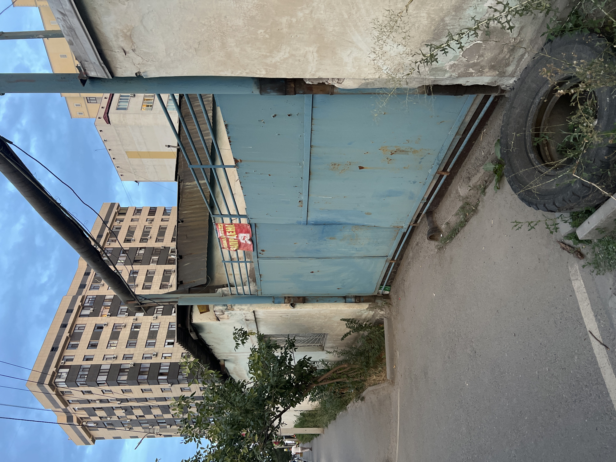 This is a street-level, eye-level, medium shot of a section of an urban area in Bishkek, Kyrgyzstan. The scene is dominated by a large, light blue, metal gate, which appears to be rusted in places. To the left of the gate, there is a narrow, paved walkway alongside a narrow road. A small, leafy tree grows on the left side of the walkway. Behind the walkway and tree, a weathered, pale blue building with a corrugated metal roof is visible. Further in the background, multiple apartment buildings of varying heights and architectural styles are seen against a cloudy, blue sky. One tall building on the left is beige with dark vertical and horizontal elements. Another building in the center is pale yellow and white, and a third building to its right is mostly pale yellow. A large, dark pipe runs diagonally across the upper portion of the frame, from the left to the right, partially obscuring the sky and buildings. A red sign with white lettering is attached to the top of the gate. In the foreground, to the right of the gate and in front of a weathered, beige wall, a discarded tire lies on the ground amidst sparse vegetation. The ground in front of the gate is dirt and gravel. A person is walking away from the camera on the walkway to the far left of the image.