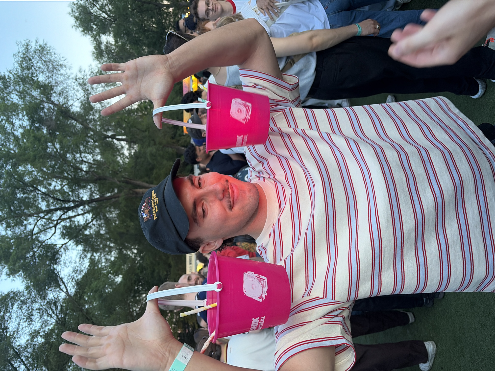 In this medium shot, a young man with a mustache, wearing a striped t-shirt and a black baseball cap, stands in what appears to be a crowded outdoor event, likely a festival, given the presence of many people and trees in the background. He is holding two bright pink buckets, each with a white handle, by their straps. Both buckets have a graphic of a turntable with a record on it. The man is looking off to the right, with his arms raised and hands spread open, as if presenting the buckets. One bucket has two straws visible inside. The man has a wristband on his left arm. The environment suggests an urban setting in Montreal, Canada, possibly Parc Jean-Drapeau during the Piknic Électronik festival, an outdoor electronic music event known for its summer series in Montreal. The lighting and time of day suggest late afternoon or early evening.