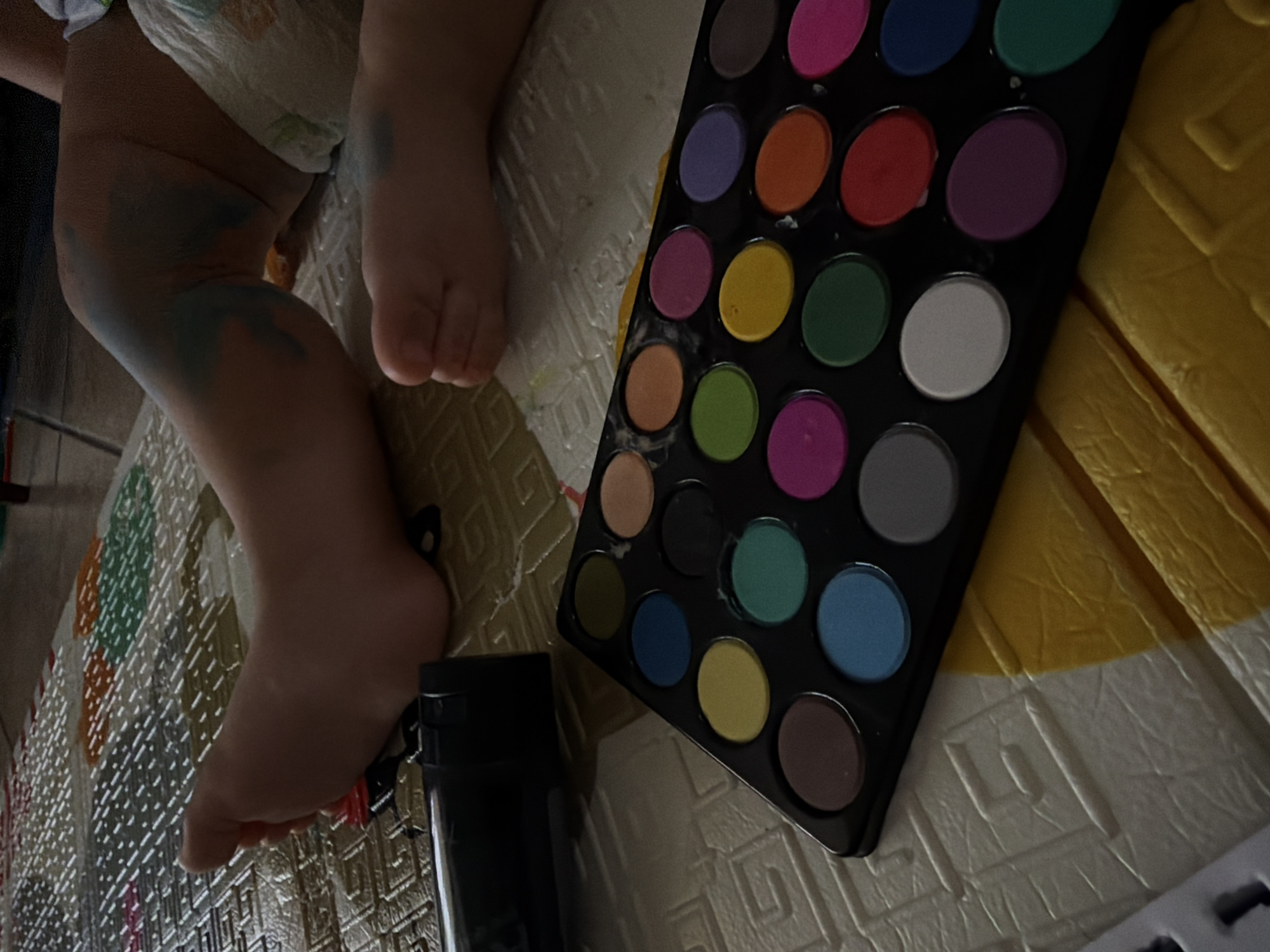 The image shows a close-up of a child's legs and feet on a mat. The child's knees have blue marks on them. A palette of colorful makeup is positioned in the foreground. The setting appears to be indoors, likely a home environment, with a mat that has a patterned surface. There is no indication of the city of Sharm El Sheikh, Egypt, or any other specific geographic location.