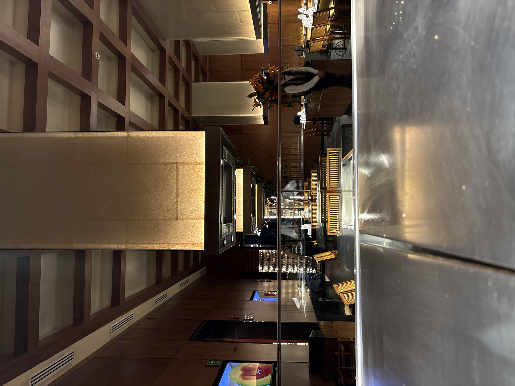 The image shows the interior of a restaurant, with a modern design featuring wood paneling, neutral tones, and metallic accents. Several people, presumably staff and customers, are visible, though their faces are not distinct. Tables and chairs are arranged throughout the space, and decorative flower arrangements add touches of color. The presence of screens displaying images suggests a contemporary atmosphere. The location of this scene in Puebla, Mexico, cannot be determined solely from this image.