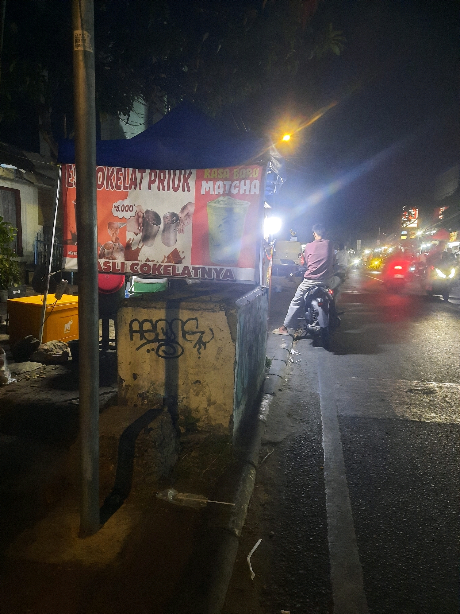 The image depicts a nighttime street scene in North Jakarta, Indonesia. In the foreground, a food stall with a blue awning displays a large banner advertising "ES COKELAT PRIUK" and "RASA BARU MATCHA." The banner includes images of drinks and a price of "8.000." To the left of the stall, a yellow cooler box is visible. A person is seated on a motorbike in the middle of the road, facing away from the camera. The road is paved and has a white line marking. In the background, there are more street lights, traffic lights, and vehicles, indicating a busy urban environment. The overall atmosphere is that of a typical street scene at night in a city.