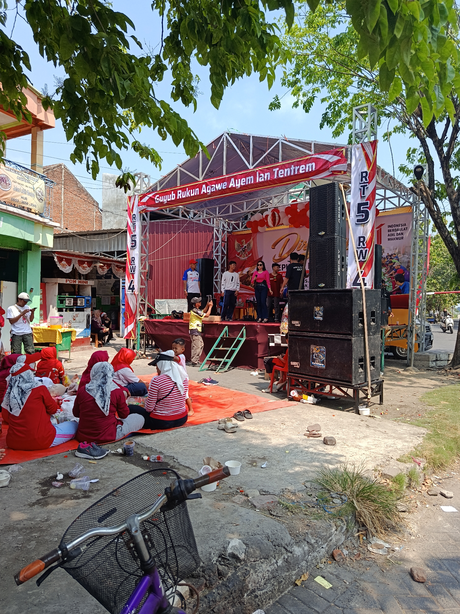 The image depicts an outdoor event with a stage set up on a street in Keboansikep, Indonesia. A crowd of people, many wearing red and white attire, are seated on the ground on an orange mat, facing the stage. Some are eating or interacting amongst themselves. On the stage, several individuals are performing, including singers and musicians. Large speakers are positioned on either side of the stage. Banners with Indonesian text are visible, including one that reads "Guyub Rukun Agawe Ayem Ian Tentrem." A purple bicycle with a basket is in the foreground. The environment appears to be a residential or commercial street with buildings and trees surrounding the event area. The overall scene suggests a community gathering or celebration.