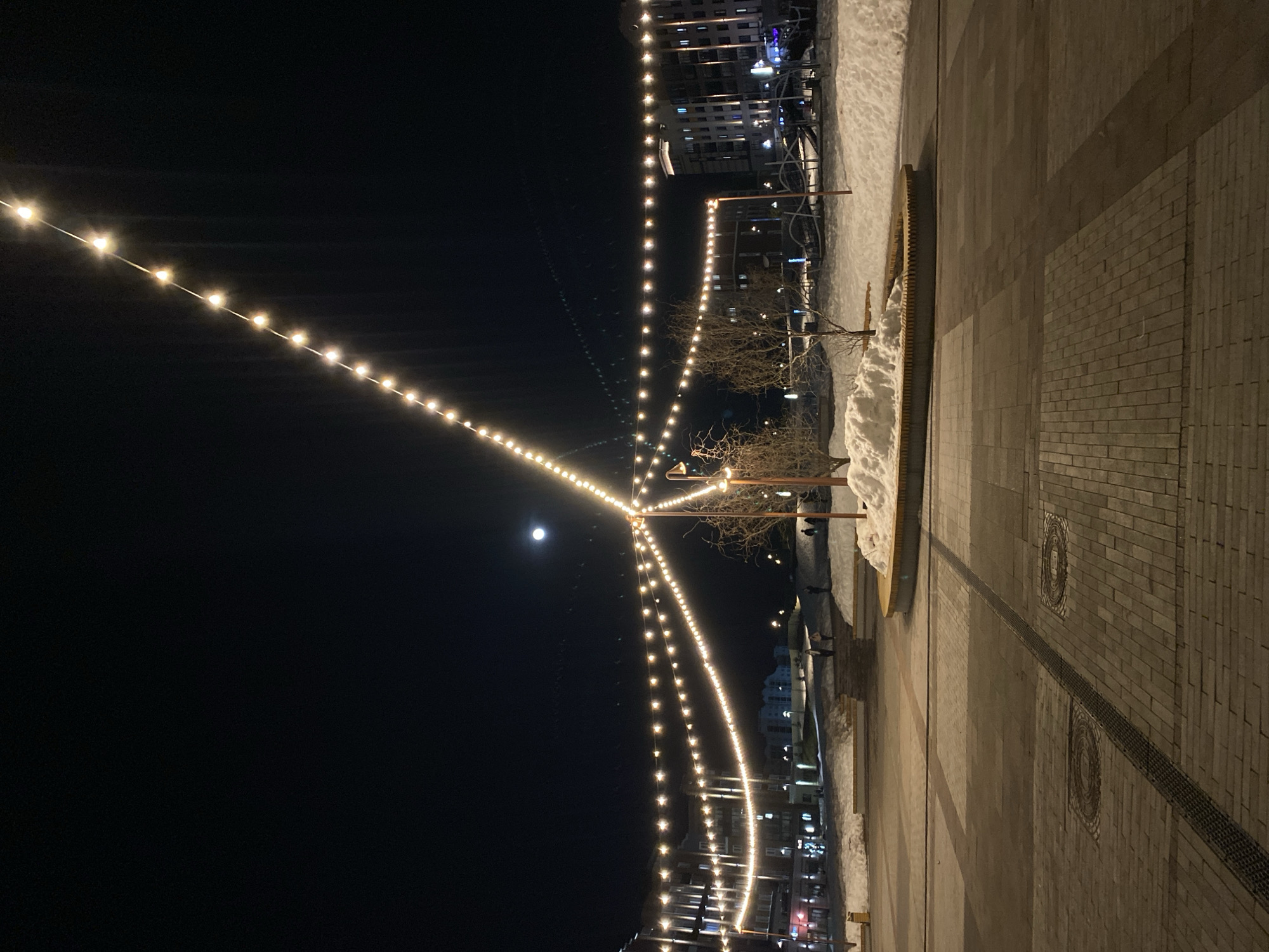 The image shows a nighttime scene in Izhevsk, Russia. Strings of lights illuminate a pedestrian area. To the right, the paved walkway is visible, with snow piled up on the edges. In the background, buildings are lit, their lights contrasting with the dark sky. The moon is visible in the sky. Some trees are visible. Several people are near the buildings, walking.