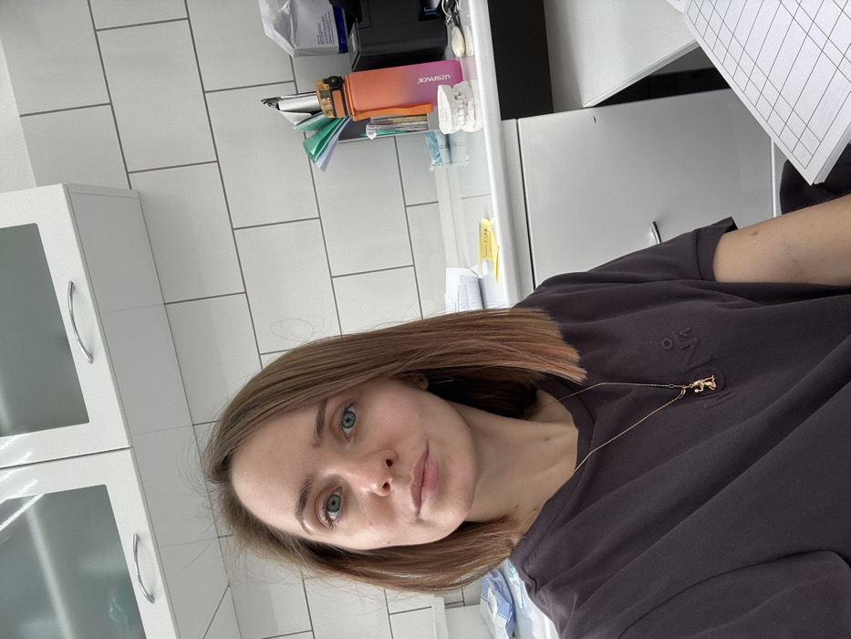 A woman with short blonde hair and blue eyes poses indoors. She wears a dark t-shirt with embossed writing and a delicate gold necklace featuring a figure. In the background, white cabinets with metal handles and a white tiled wall are visible. A desk with assorted items, including an orange water bottle and dental molds, is also present.