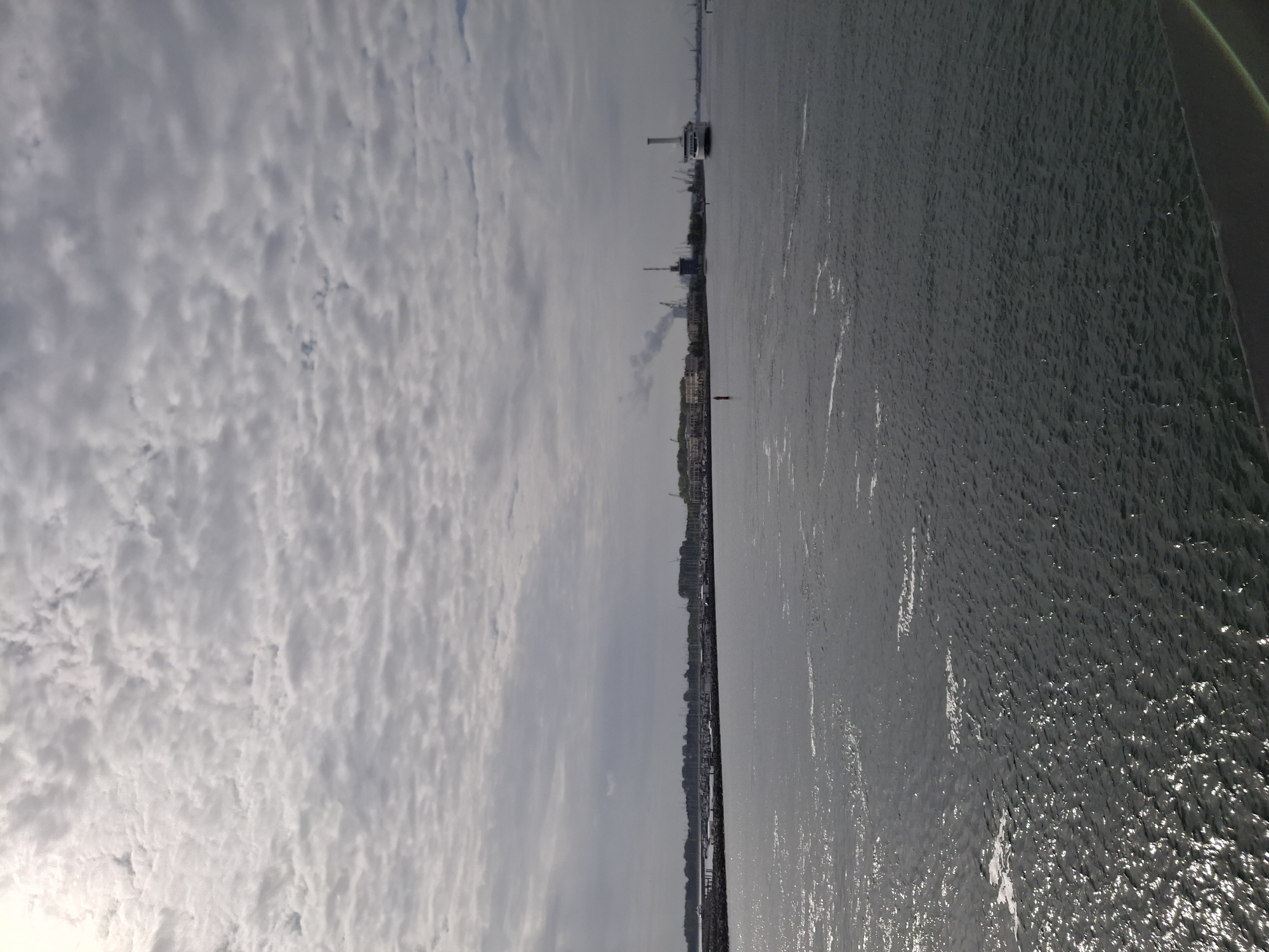 The image captures a view of the Warnow River in Rostock, Germany, under a sky filled with textured clouds. The river occupies the right half of the image and shows the surface rippled by waves. Along the riverbank on the right side, a row of buildings, trees, and possibly marina structures are visible in the distance. A red buoy is located in the water near the shore. In the far distance, a ferry or ship can be seen on the river.