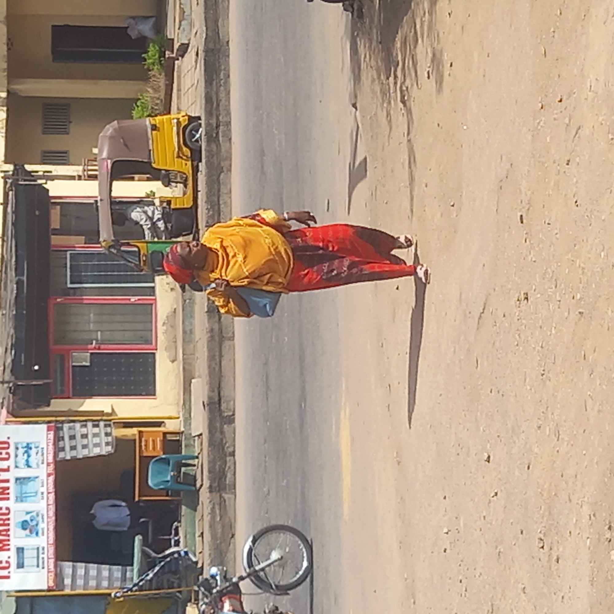 The image depicts a street scene in Dadi Riba, Nigeria. A woman wearing a yellow top and red patterned trousers is walking along the road, carrying a blue bag. A yellow auto-rickshaw is parked on the side of the road, with a person visible inside. A motorcycle is parked near a building with a sign reading "I.C. MARC INTL CO." A blue chair is placed near the building entrance. The street appears dusty and the buildings are of modest construction.