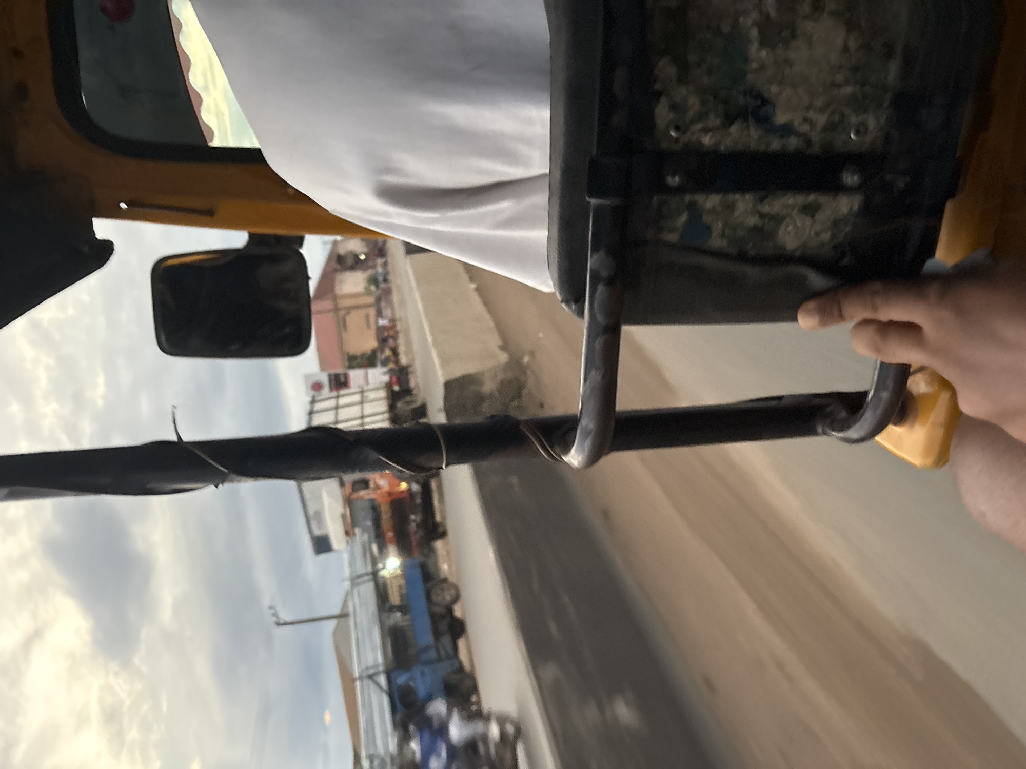 The image is a first-person perspective from within a yellow auto-rickshaw, likely in Onitsha, Nigeria, moving along a dusty road. The sky is overcast with dramatic clouds. In the foreground, a person's hand is visible, touching the side of the auto-rickshaw. The interior of the auto-rickshaw is visible, including a dark seat with a patterned insert and a metal bar. Outside the vehicle, several other vehicles are present, including a blue truck loaded with silver metal sheets, an orange truck, and a motorcycle with a rider. Buildings and shops line the side of the road, and a concrete barrier separates the road from the area where the buildings are situated. The overall scene depicts a busy urban or semi-urban environment during daylight hours, possibly late afternoon due to the lighting.