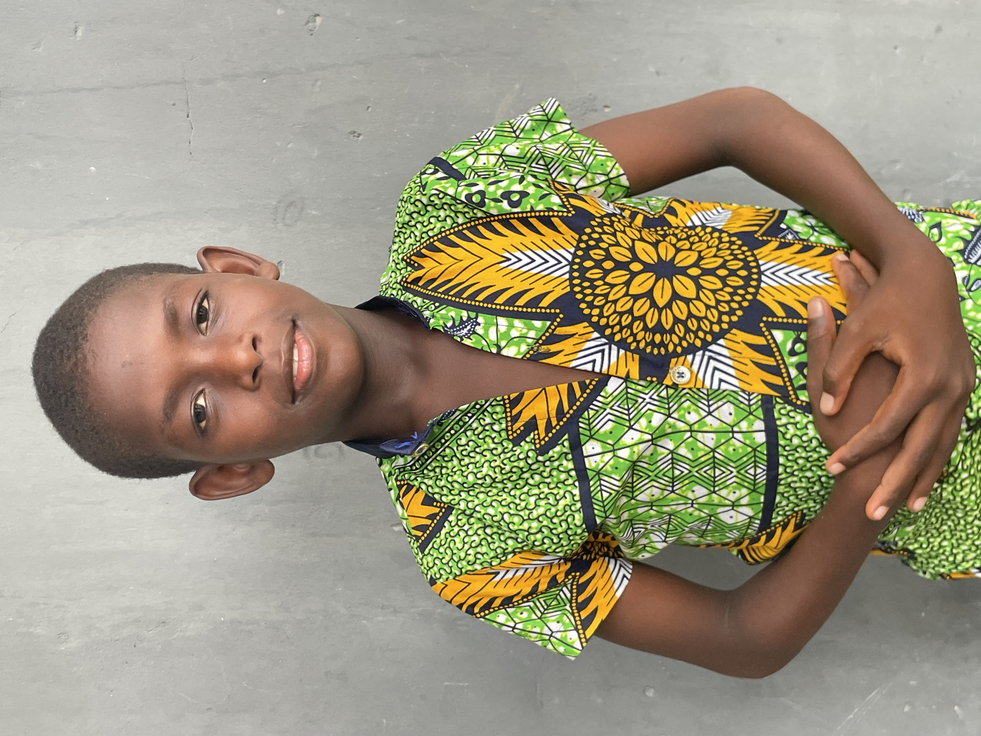 A young boy with dark skin and short, dark hair lies on a light grey concrete surface, looking directly at the camera with a gentle smile. The image is oriented such that he appears to be lying on his left side, with his head slightly raised. He is wearing a vibrant, short-sleeved shirt featuring a bold pattern of bright green, yellow, black, and white. A prominent, sunflower-like motif in yellow and black is centered on his chest, surrounded by various geometric and abstract designs. His left hand rests on his midsection, while his right arm is bent with his hand also near his body. The concrete surface beneath him shows minor cracks and texture. The lighting is bright and even, suggesting it is daytime. The boy's expression conveys a sense of calm and approachability. This scene likely takes place outdoors or in a simple, unadorned indoor space in Badagry, Nigeria. There is no visible text.