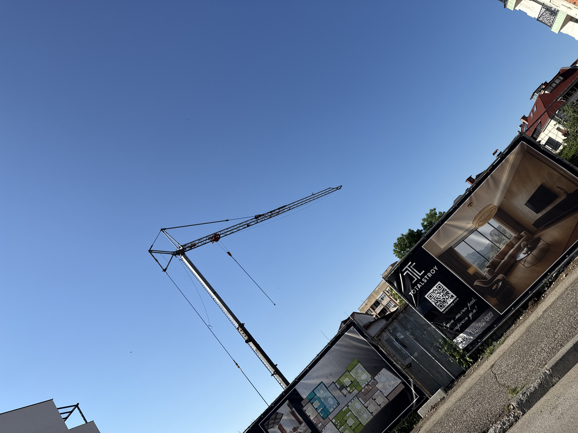 An upward-angle, wide shot shows a construction site with a tall crane against a clear blue sky. A banner on the left displays floor plans for an apartment complex. Another banner to the right, featuring the logo "TOTALSTROY", includes a QR code, a website address, a phone number, and an image of a modern living room. In the background, buildings are visible, some under construction and others already completed. The foreground is a paved area, possibly a sidewalk or road. The image captures a sunny day in Vratsa, Bulgaria.