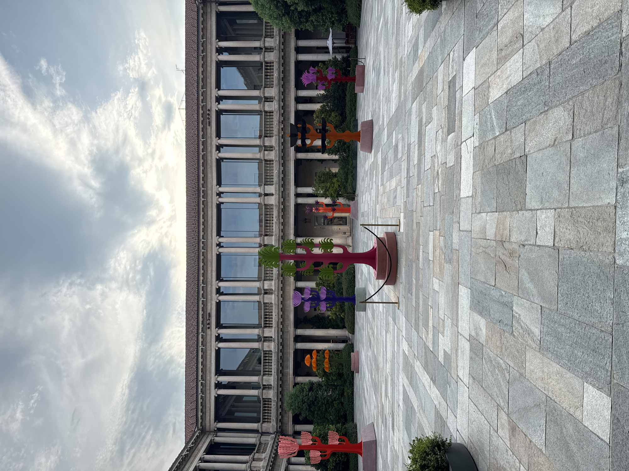 The image depicts the courtyard of a building in Milan, Italy, likely the Palazzo delle Stelline, given the architectural style and courtyard layout. The courtyard is paved with large, rectangular stone tiles in varying shades of grey and beige. In the center of the courtyard are several brightly colored, stylized tree sculptures. These sculptures are abstract, with layered, fan-like branches and are rendered in vibrant hues of red, orange, purple, and green. They stand on square bases. The background features a grand, classical building with a colonnade of tall, grey columns supporting an upper story with large, reflective windows. Greenery, including trees and bushes, is visible behind the sculptures and along the sides of the courtyard. The sky above is overcast with scattered clouds. There are no people visible in the image.