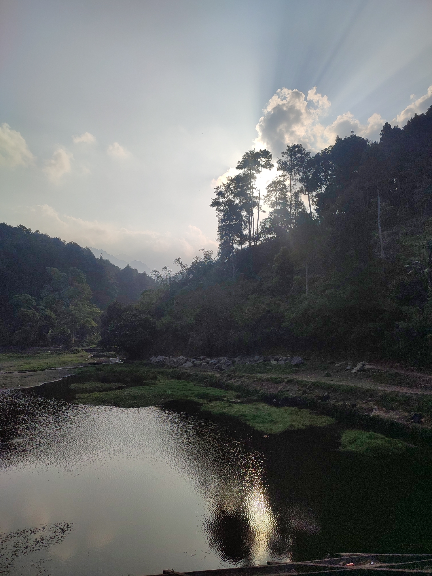 The image depicts a scenic view of a tranquil body of water surrounded by lush, verdant hills and dense foliage. The sun is setting, casting a warm glow and creating a dramatic interplay of light and shadow. Sunbeams pierce through the clouds, illuminating the sky with a celestial radiance. The water's surface is calm, reflecting the surrounding landscape and the ethereal light.
This is the location of a waterfall in Joho, Indonesia. The area is characterized by its natural beauty and offers a peaceful retreat from the hustle and bustle of urban life. The image captures the serene ambiance of this natural wonder, inviting viewers to immerse themselves in its tranquil embrace.