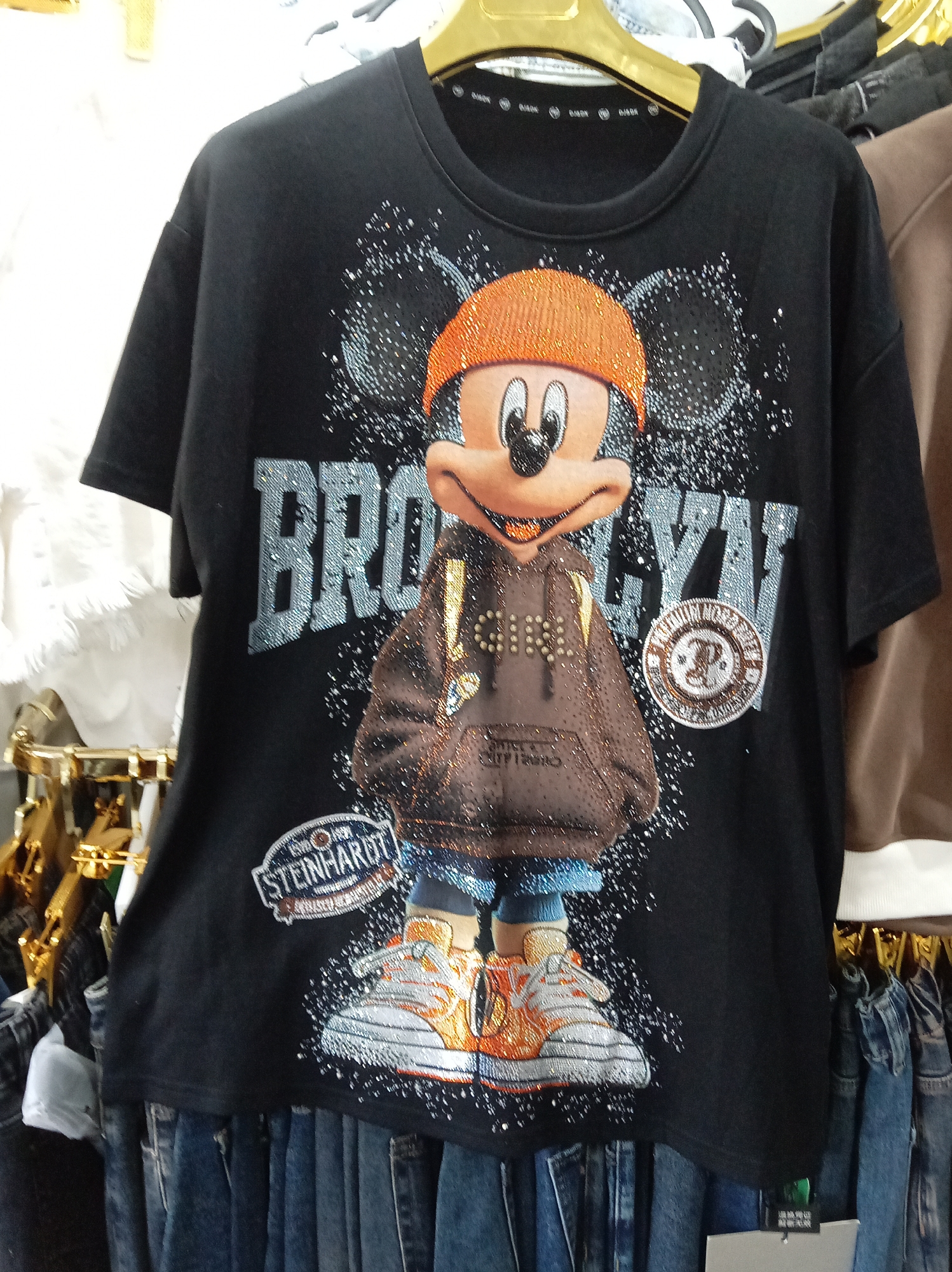 A black t-shirt with a sequined image of Mickey Mouse wearing a hoodie and sneakers is displayed on a hanger in a retail environment. The t-shirt features the word "BROOKLYN" in large, sparkly letters behind Mickey Mouse, and a circular patch with the word "STEINHARDT" on the lower left. The background includes racks of clothing and other retail fixtures. The image is an indoor shot of clothing merchandise. There is no indication that this image was taken in Donetsk, Ukraine.