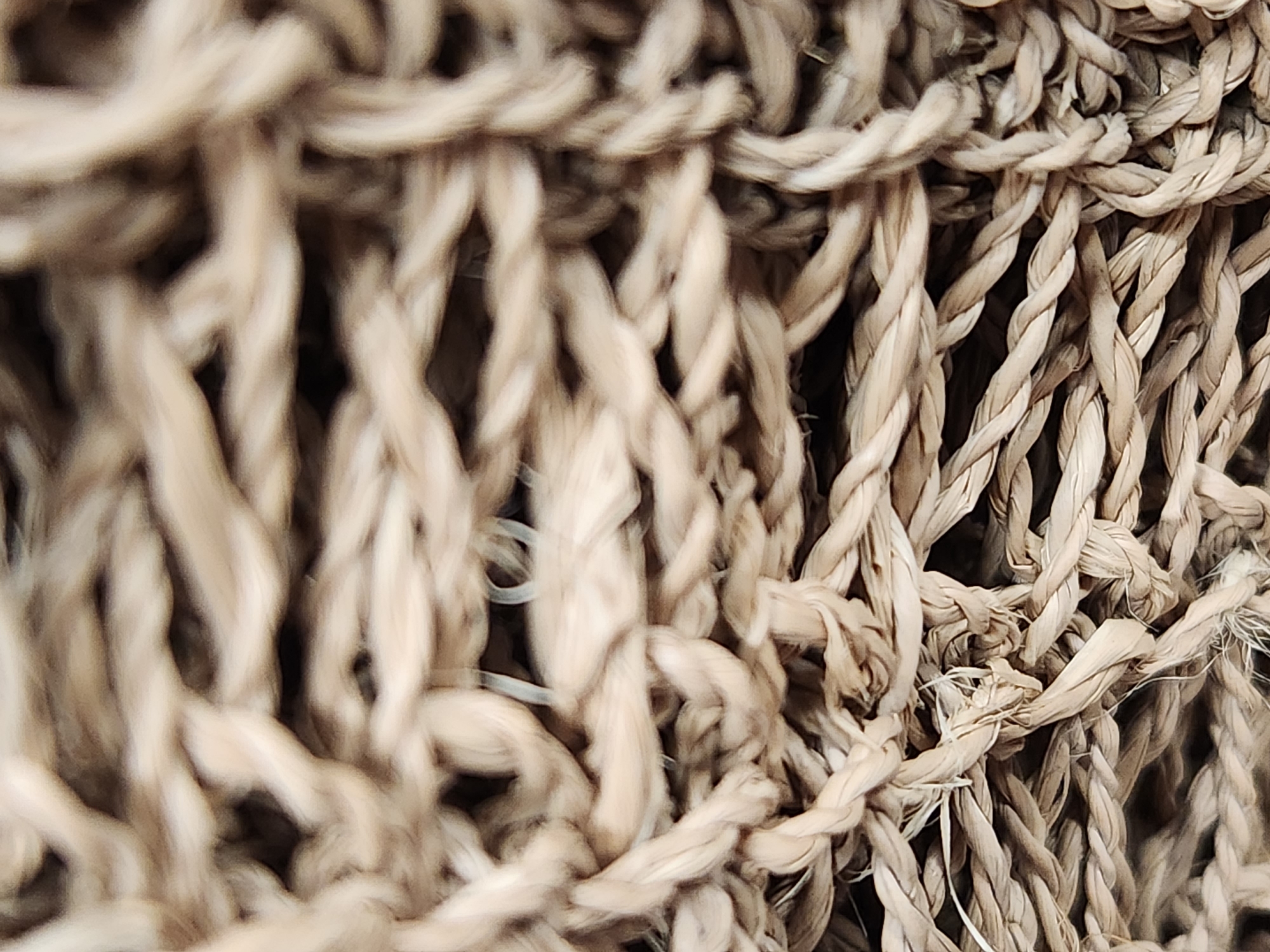 The image is a close-up, abstract shot of tightly woven rope. The rope is a light brown, natural fiber material, possibly jute or hemp, with visible strands twisted together to form thicker cords. The texture is rough and fibrous, with some individual fibers appearing loose and frayed, especially in the lower half of the image. The lighting is somewhat dim, creating shadows between the ropes, which enhances the sense of depth and texture. The focus is shallow, with the foreground ropes being slightly sharper than those in the background, which are considerably blurred. There are no people, animals, or discernible objects other than the rope itself. The image does not depict any recognizable landmarks or features that would allow for its specific location within Maitland, United States of America, or any other city. The scene is purely a textural study of the rope material.