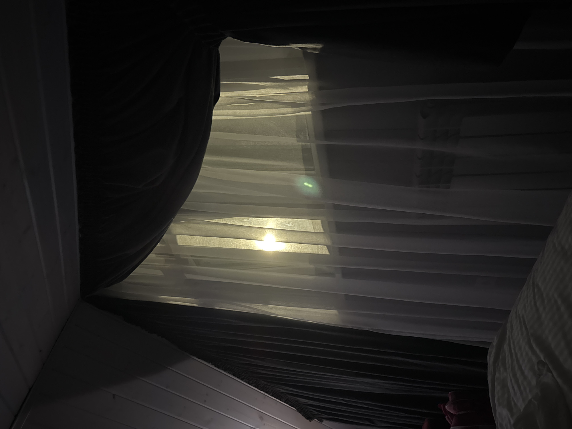 The image depicts an interior scene, likely a bedroom, illuminated by a soft, warm light emanating from a window covered by sheer white curtains. The curtains are drawn, and the light appears to be from an external source, possibly the sun or artificial lighting, creating a diffused glow. Behind the sheer curtains, a darker, heavier curtain is visible, suggesting a layered window treatment. The ceiling appears to be sloped and paneled with wood, common in attic or dormer rooms. A radiator is faintly visible behind the sheer curtains. In the foreground, the corner of a bed with striped bedding is visible, indicating the vantage point of the photograph. The overall ambiance is serene and dimly lit, with a focus on the interplay of light and fabric. No people or specific actions are depicted. The location information provided, "Doma 1096 Km, Russia," is too vague to provide specific environmental context beyond a general indication of a residential setting in Russia.