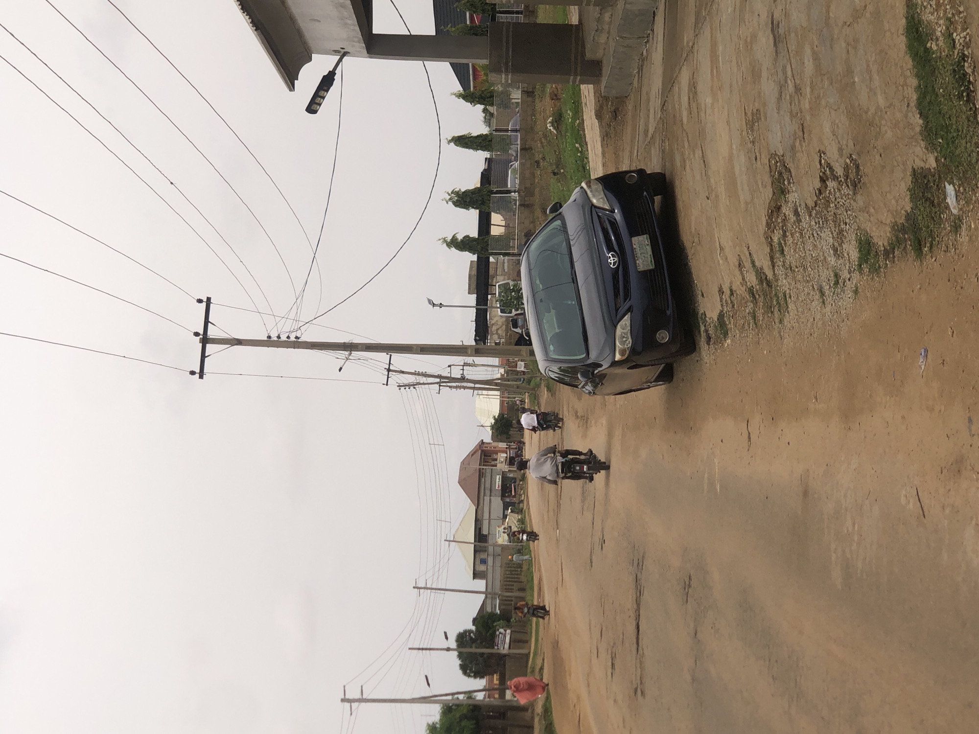 The scene depicts a street in Hanwa, Nigeria, on what appears to be an overcast day. A dark blue car is parked on the right side of the road, partially obstructing the roadway. Several motorcycles are moving down the street in the same direction as the parked car. Buildings and vegetation line the street. Utility poles and overhead wires dominate the sky. The road is unpaved and has a light brown/beige color.