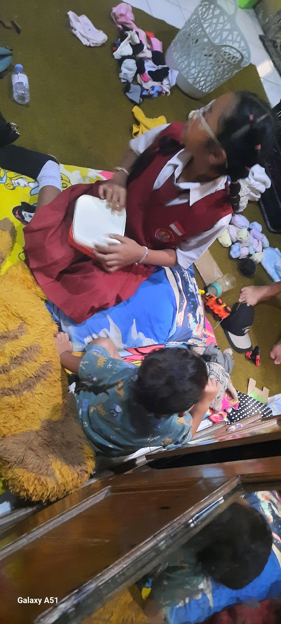 The image depicts an indoor scene, possibly a living room or bedroom, with children present and scattered items on the floor. Two children are the primary focus. In the foreground, a younger child, wearing a green and yellow patterned t-shirt, is seated on a blue and white patterned mat. They are looking down. To their left, a fluffy yellow mat with brown stripes is visible.
Further back, an older child, wearing a maroon and white school uniform, is also seated. They are holding a white rectangular object, possibly a lunchbox or a tray, in their hands and appear to be interacting with it. The older child is wearing glasses and has their hair in braids.
The environment is cluttered with various items. A pile of colorful socks and clothing is near a light-colored laundry basket. A plastic water bottle is on the floor to the left. Other items scattered around include a black and gray baseball cap, a small red toy truck, and what appears to be a cardboard box. The flooring is a dark green carpet. The background shows a wooden surface, possibly furniture.
The overall impression is one of a casual, possibly domestic setting, with children engaged in activities on the floor amidst household items. The context of "Jatitengah, Indonesia" suggests this is a scene from that specific location.