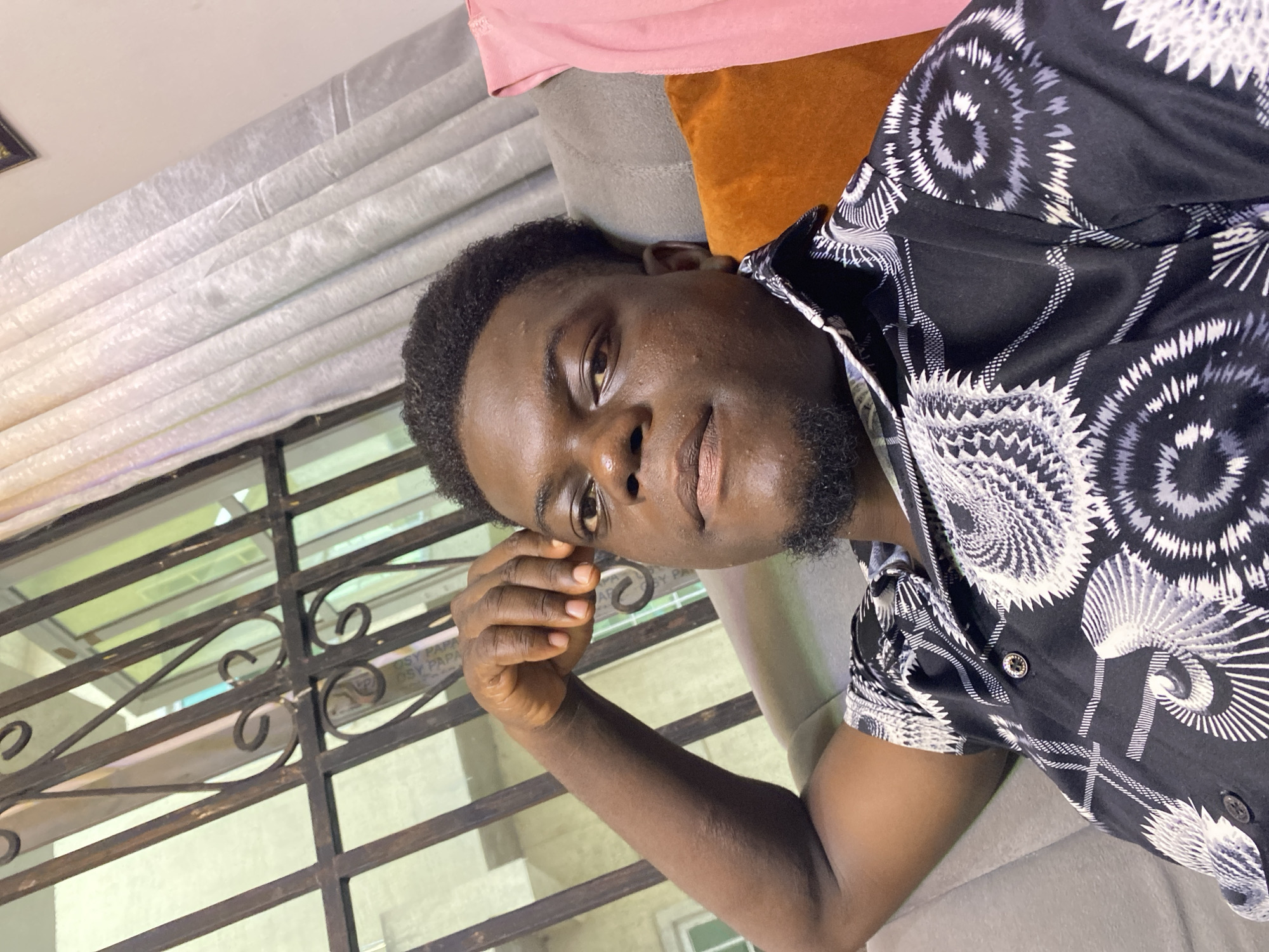 A close-up, slightly angled, portrait shot of a dark-skinned man, possibly in his late teens or early twenties. He has short, dark hair, a trimmed beard, and is wearing a patterned, short-sleeved shirt with what appear to be floral or starburst designs in black and white. The setting appears to be indoors, with a window behind the man, featuring a decorative metal grill or bars. Outside the window, there are blurry hints of greenery and possibly other buildings. The man is leaning against a grey cushion, with hints of a pink fabric above and an orange cushion behind. The location is Unknown, Unknown. The man's right hand is raised near his face, with his fingers lightly touching his temple. He is looking directly at the camera with a relaxed expression. The lighting appears to be soft and diffused.