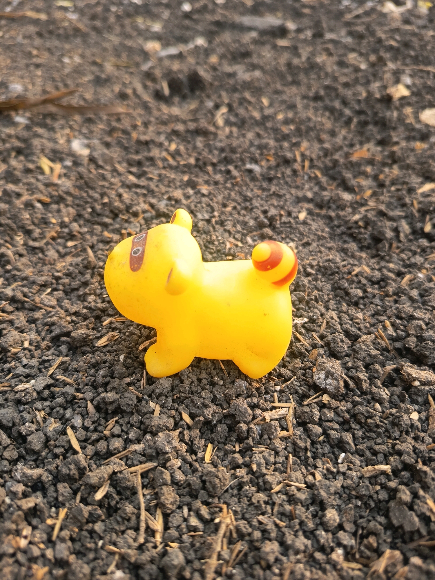 A yellow toy dog with red markings on its ears and tail lies on dark, crumbly soil. The soil contains small pebbles and dried plant matter. The lighting suggests it is outdoors during the daytime. The toy is positioned in the center of the frame, facing left, with its body parallel to the ground. The background is a blurred expanse of the same soil. There is no indication of human presence or any specific location within Sidorejo, Indonesia, beyond it being an outdoor environment.