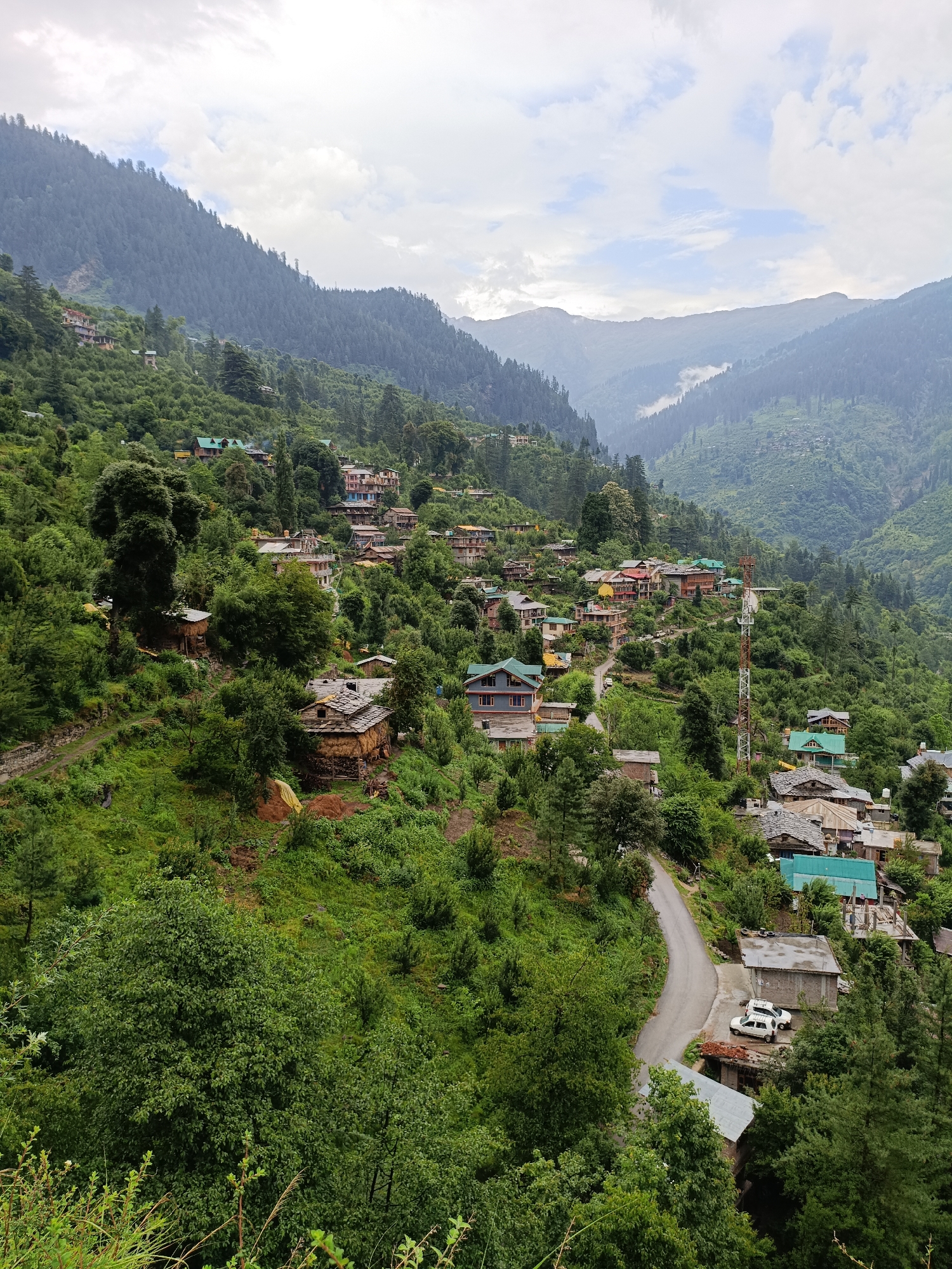 This is a high-angle, medium shot of a village nestled in a lush green mountain valley. The village, identified as Damadi, India, is characterized by traditional stone and wooden houses with slate or tin roofs, interspersed with more modern structures. A winding paved road cuts through the scene, with two white cars parked near a building on the right side of the road. The houses are scattered across the steep, verdant slopes, surrounded by dense forests of pine and deciduous trees. In the background, mist-shrouded mountains rise in layers, suggesting a high-altitude location. The sky is overcast with scattered clouds. No people are visible in the frame. The overall impression is one of a remote, tranquil mountain community.