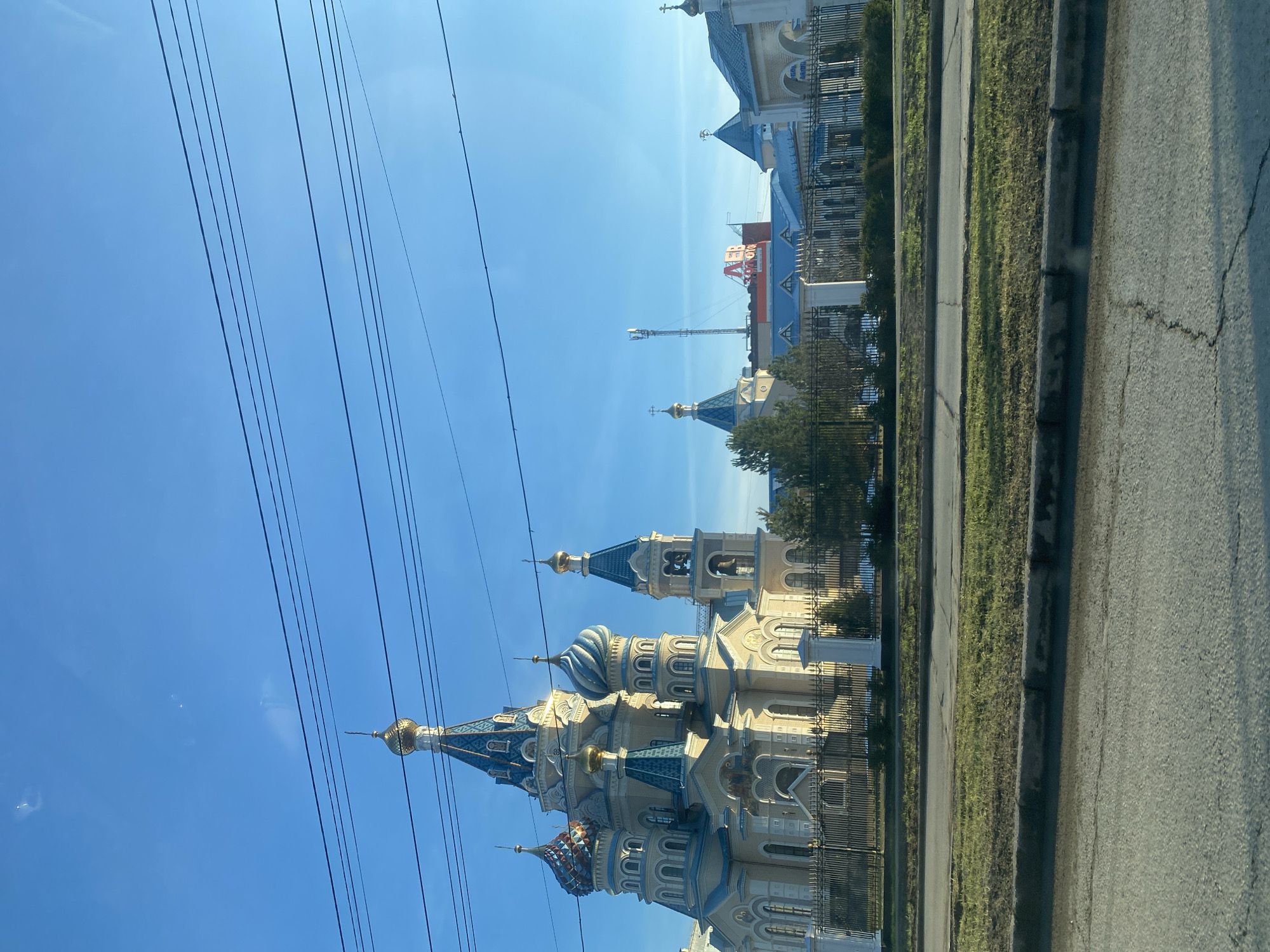 The image shows a view of the Alexander Nevsky Cathedral in Izhevsk, Russia, taken from what appears to be a moving vehicle. The cathedral, with its distinctive onion domes, is fenced off and located next to a road. Overhead power lines are visible against the clear blue sky. Other buildings are discernible in the background. The scene presents a typical urban landscape featuring a religious structure.