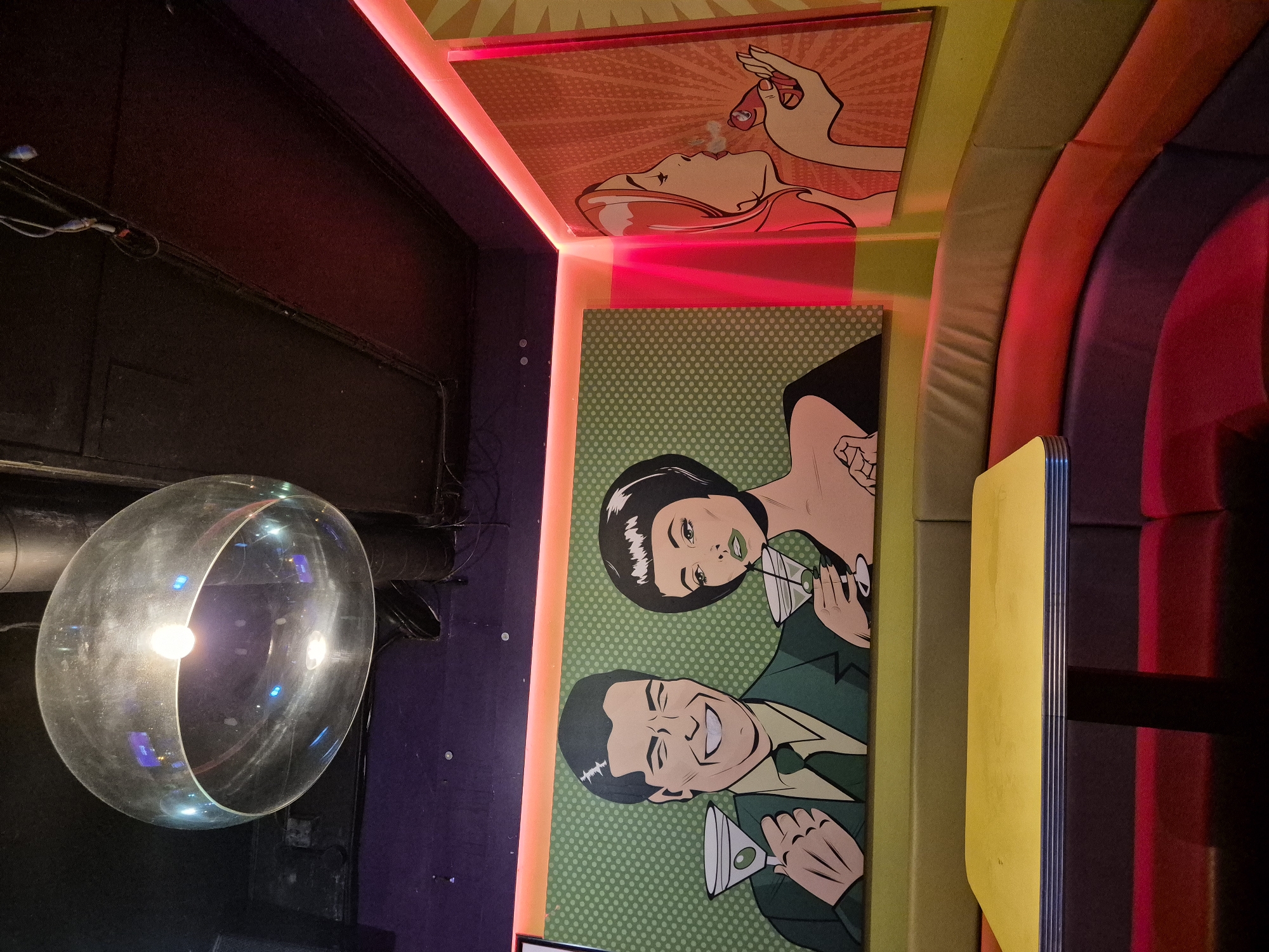 The image shows a portion of a brightly lit interior space, likely a bar or lounge, with a focus on colorful artwork and seating.
On the right is a section of curved seating, featuring alternating colors of yellow, beige, red, and purple/burgundy upholstery. On the walls are two framed, comic-book-style art pieces. The one above depicts a person smoking a cigarette or similar item, while the one below shows a man and a woman holding martini glasses against a green polka-dot background.
To the left, there is a large, spherical light fixture with a bulb inside. The background is predominantly dark, with hints of black and purple walls.
While specific details connecting it directly to Cardiff, United Kingdom are not discernible from the image alone, it appears to be a trendy or stylized venue with an emphasis on vibrant decor.