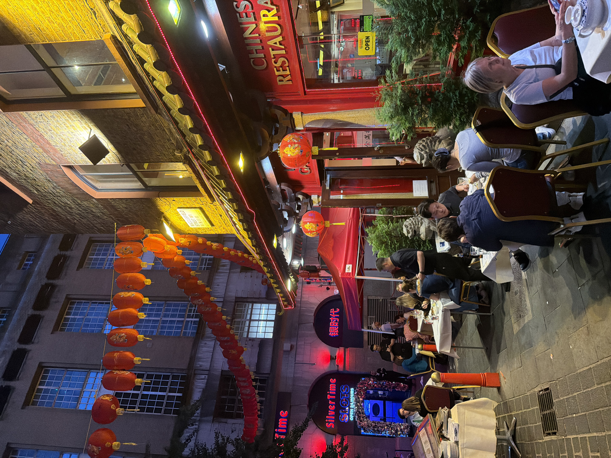The image shows a street scene in London's Chinatown, United Kingdom. It is evening, and the street is illuminated by numerous red lanterns and neon signs. A Chinese restaurant with red exterior is visible, and people are dining at tables set up outside. Lanterns hang in long strings. The architecture consists of traditional Chinese elements mixed with standard building facades. There are signs with both English and Chinese characters, including "Silver Time SLOTS" and "CHINESE RESTAURANT". People are seated at tables, apparently eating and socializing. The street is paved with cobblestones.