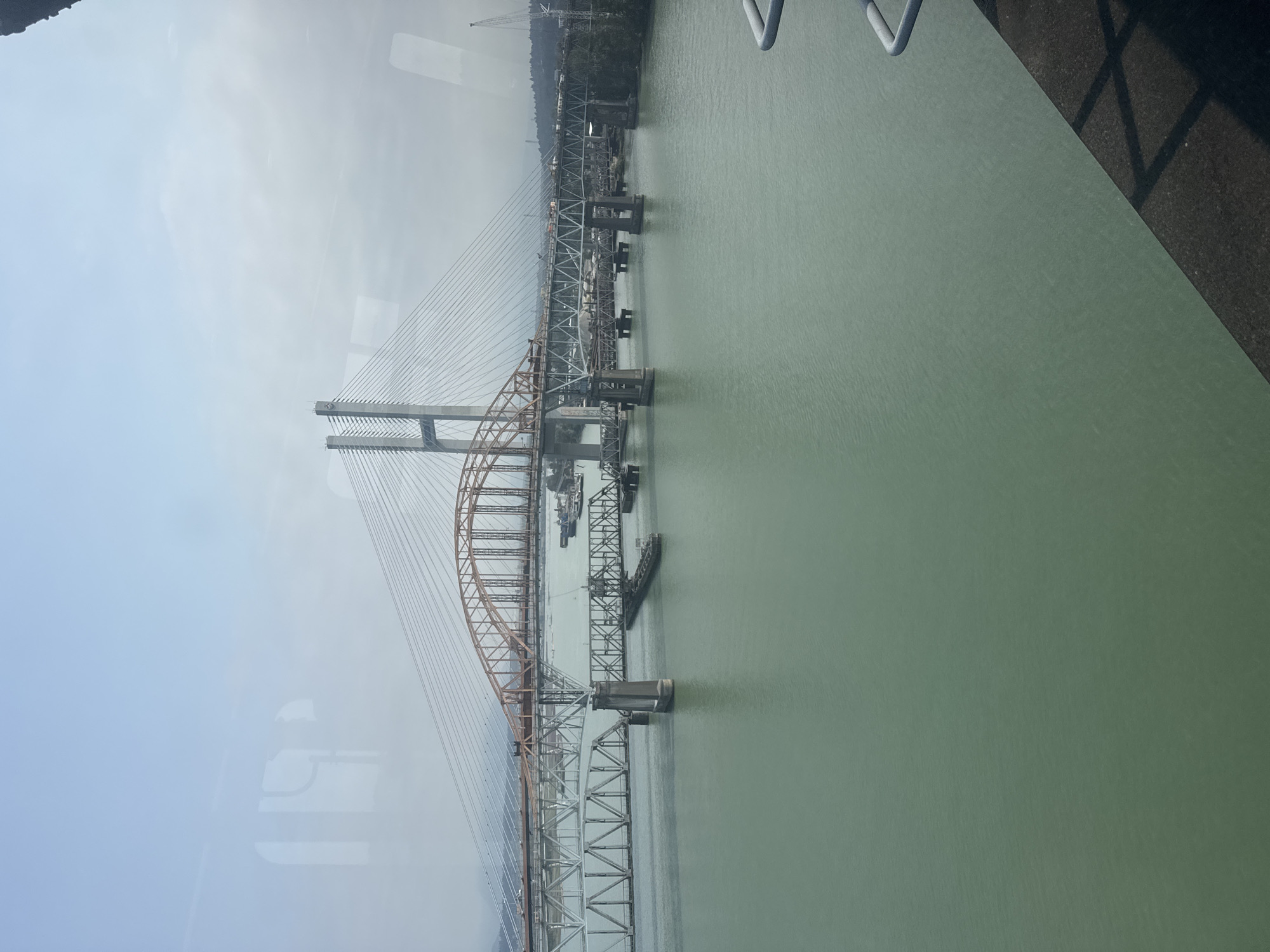 The image shows multiple bridges spanning a body of water in New Westminster, Canada. The foreground features a green, calm river or inlet, with a dark concrete embankment and railing visible in the lower right corner. The middle ground is dominated by several bridges. From left to right, there's a truss bridge with a parallel track. Behind it, a larger, modern cable-stayed bridge with tall white towers and a network of cables rises. In front of the cable-stayed bridge, a lower, orange-colored arched bridge is visible, also spanning the water. Several barges or boats are present in the water near the bridges. The background shows a hazy shoreline with trees and industrial elements, including a construction crane. The sky is overcast with a light blue hue. Reflections of a window frame are visible in the upper left portion of the image.