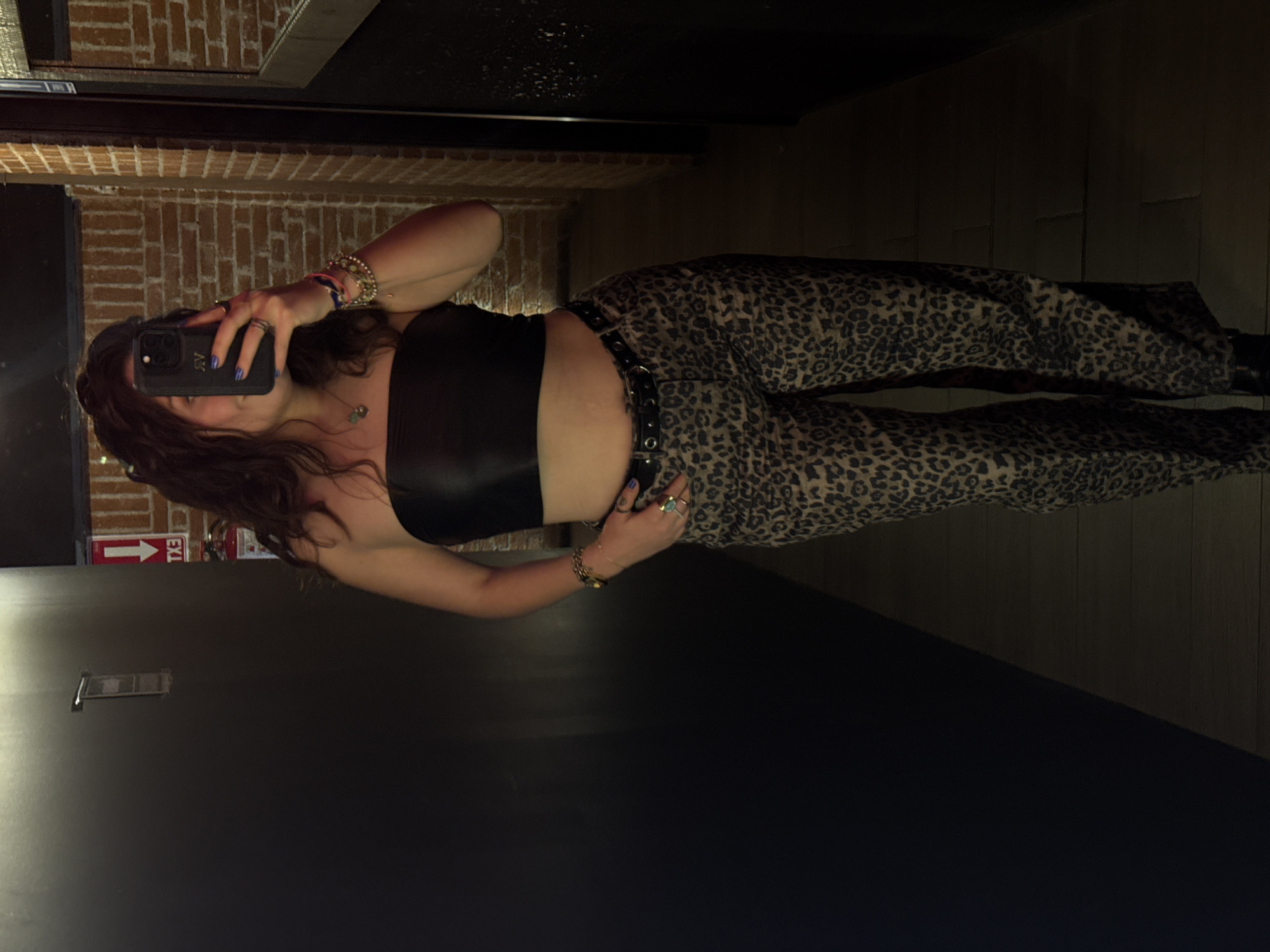 A woman is taking a selfie in a dimly lit interior space. The walls consist of brick and dark paneling, and there's a mirror reflecting the scene. An exit sign is visible. The woman is wearing a black tube top and leopard print pants with a belt. She's holding a phone in her left hand, adorned with bracelets and rings. The location, San Bernardino Tlaxcalancingo, Mexico, cannot be independently confirmed from the image.