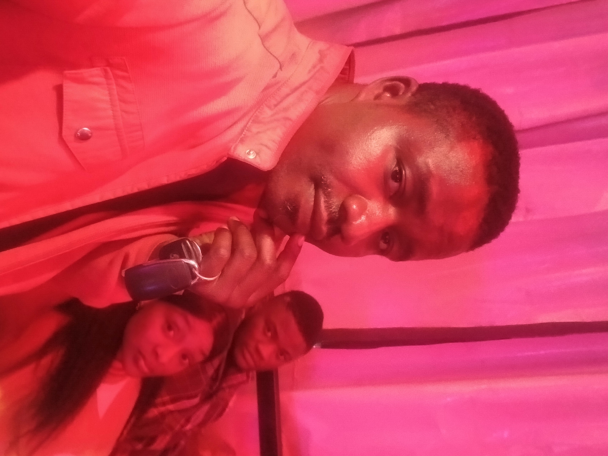 Three people are visible in a room illuminated by a red light. The man in the foreground is wearing a button-down shirt. He has short, dark hair and is holding a dark object in his hand near his face. Behind him to the left, a young woman with long, dark hair is wearing a light-colored shirt. To the right of the woman, a man with short, dark hair is partially visible. The background appears to be a wall with horizontal stripes. The location is Rukuba Road, Nigeria.